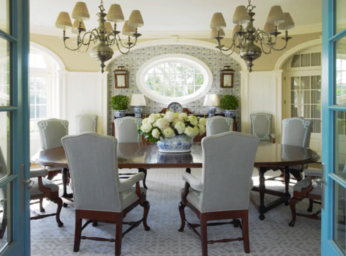 Elegant formal dining room with gray upholstered chairs, dark wood table, dual chandeliers, and a large oval window above a marble mantelpiece with white floral arrangement, designed by Pamela R Ackerman.