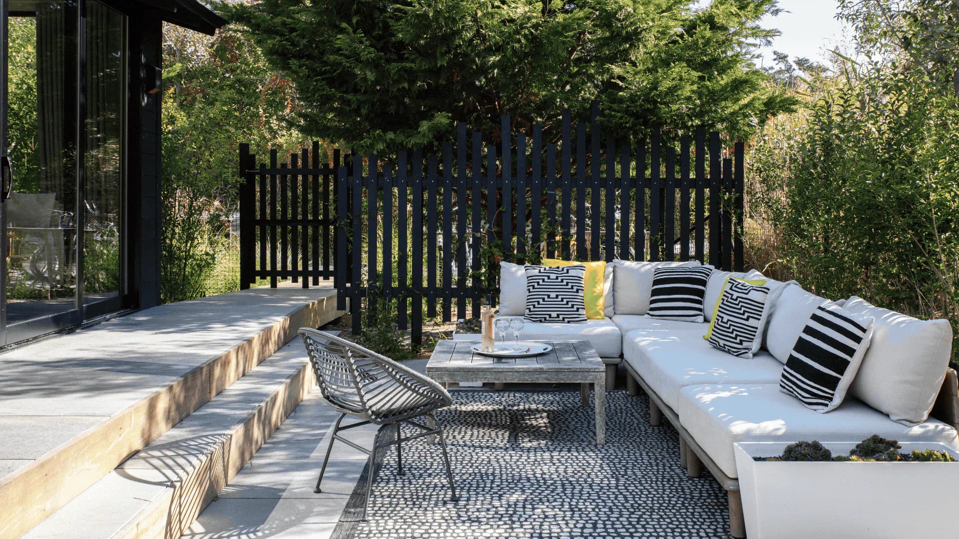 Modern outdoor patio with black slatted fence, light blue sectional sofa, patterned area rug, and metal dining chairs by Paola Reyes.