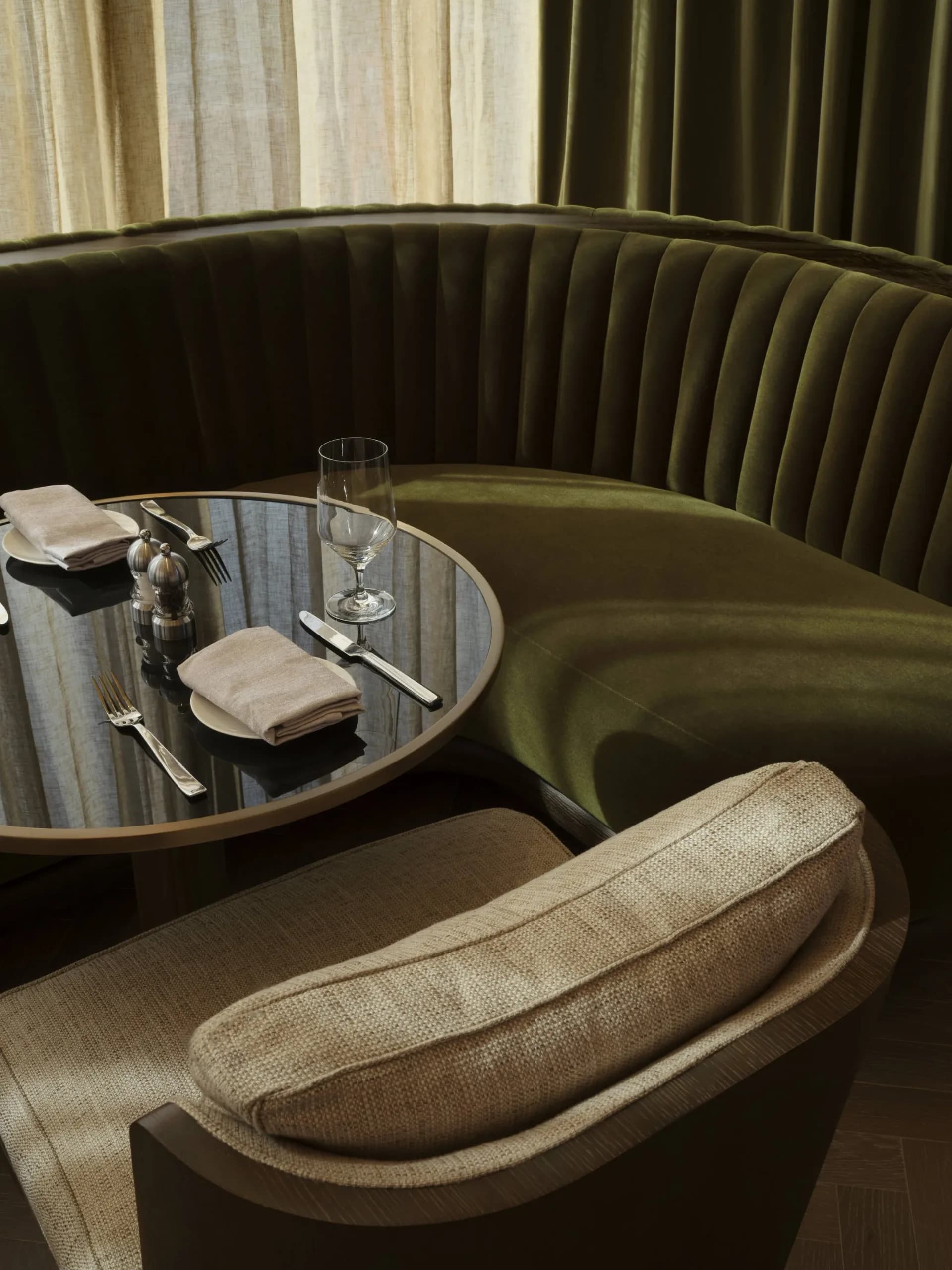 Curved banquette seating with beige upholstery, round marble table setting, and olive green pleated wall panels in an intimate dining booth by Paolo Ferrari.