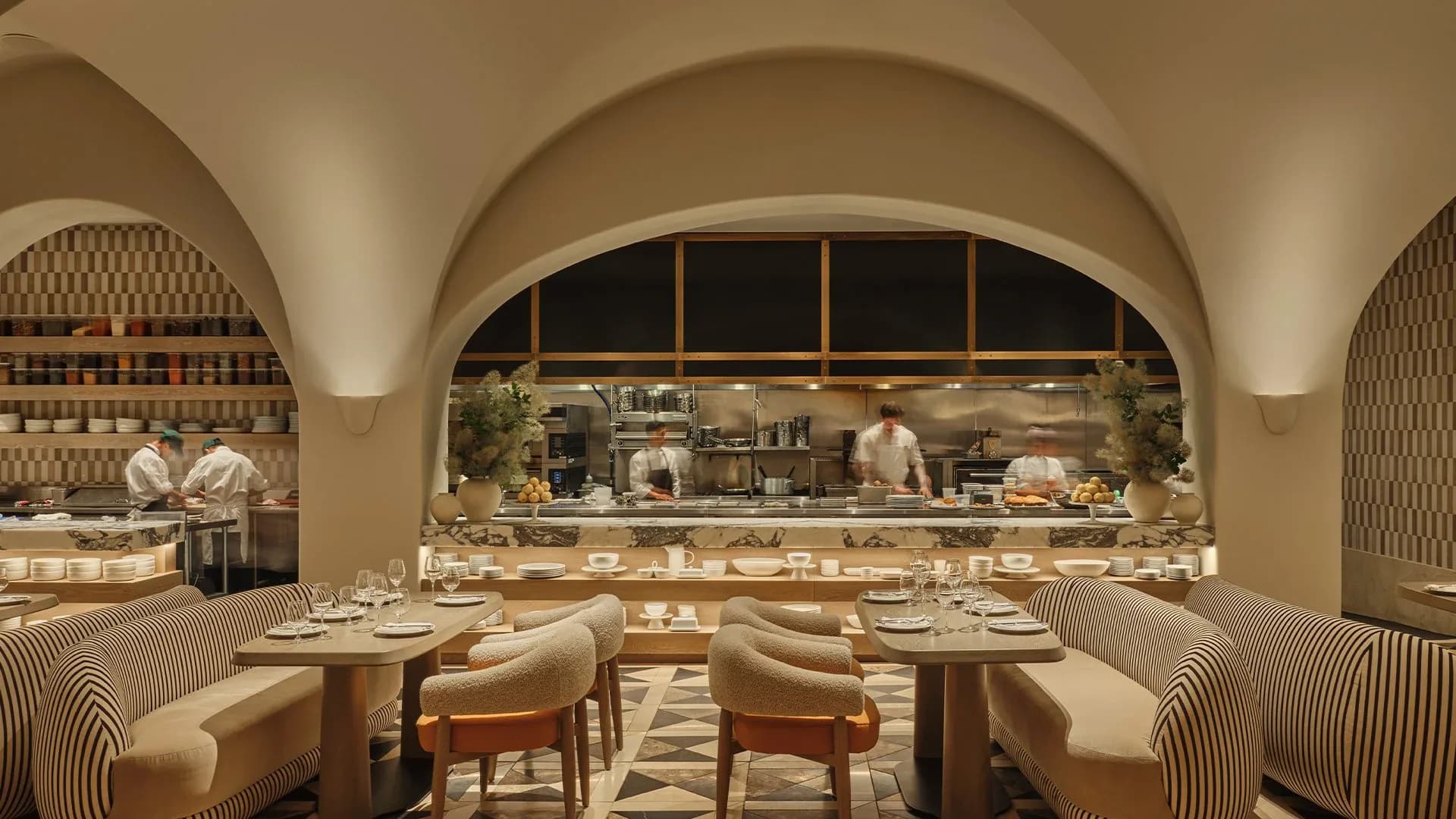 Contemporary restaurant interior with arched ceilings, striped curved banquettes, open kitchen visible through large arched window, warm neutral palette by Paolo Ferrari.