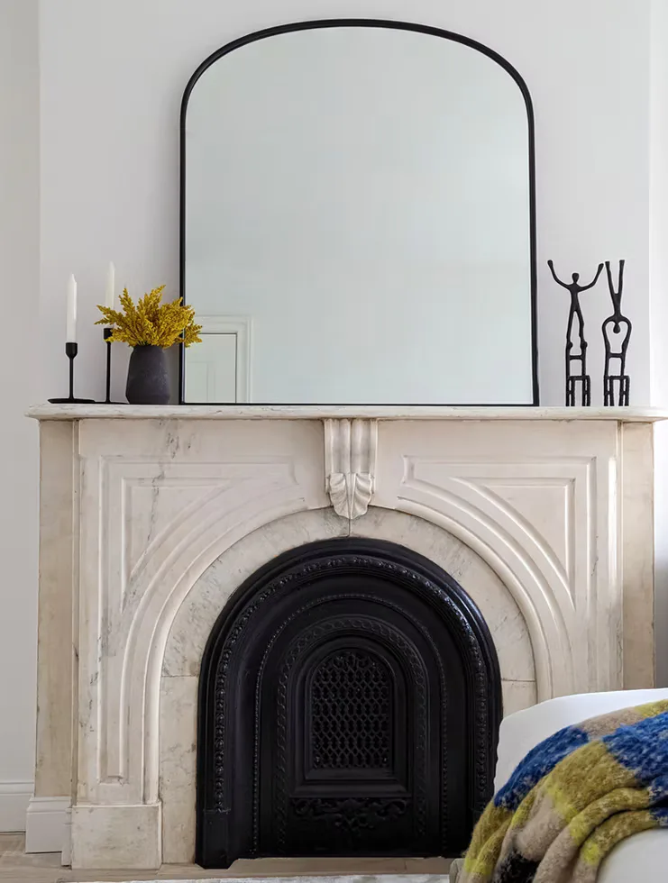 Minimalist fireplace mantel with black arched mirror, brass candlestick, potted golden flowers, and sculptural decor by Parul Ghei.