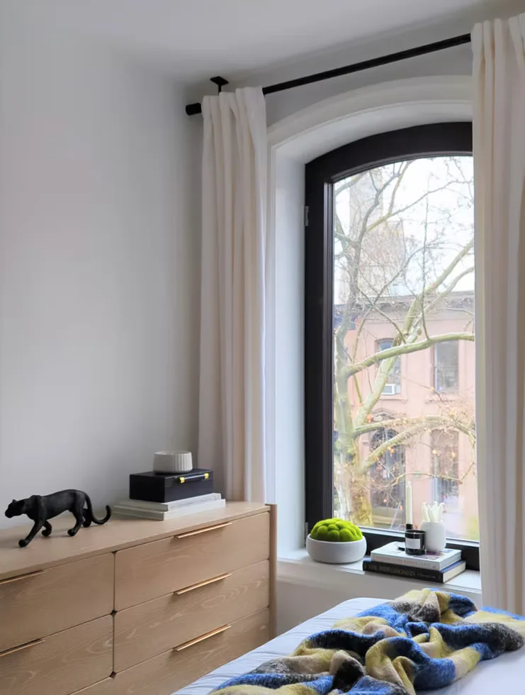 Bright modern bedroom with natural wood dresser, cream curtains, black-framed arched window, and minimalist decor by Parul Ghei.