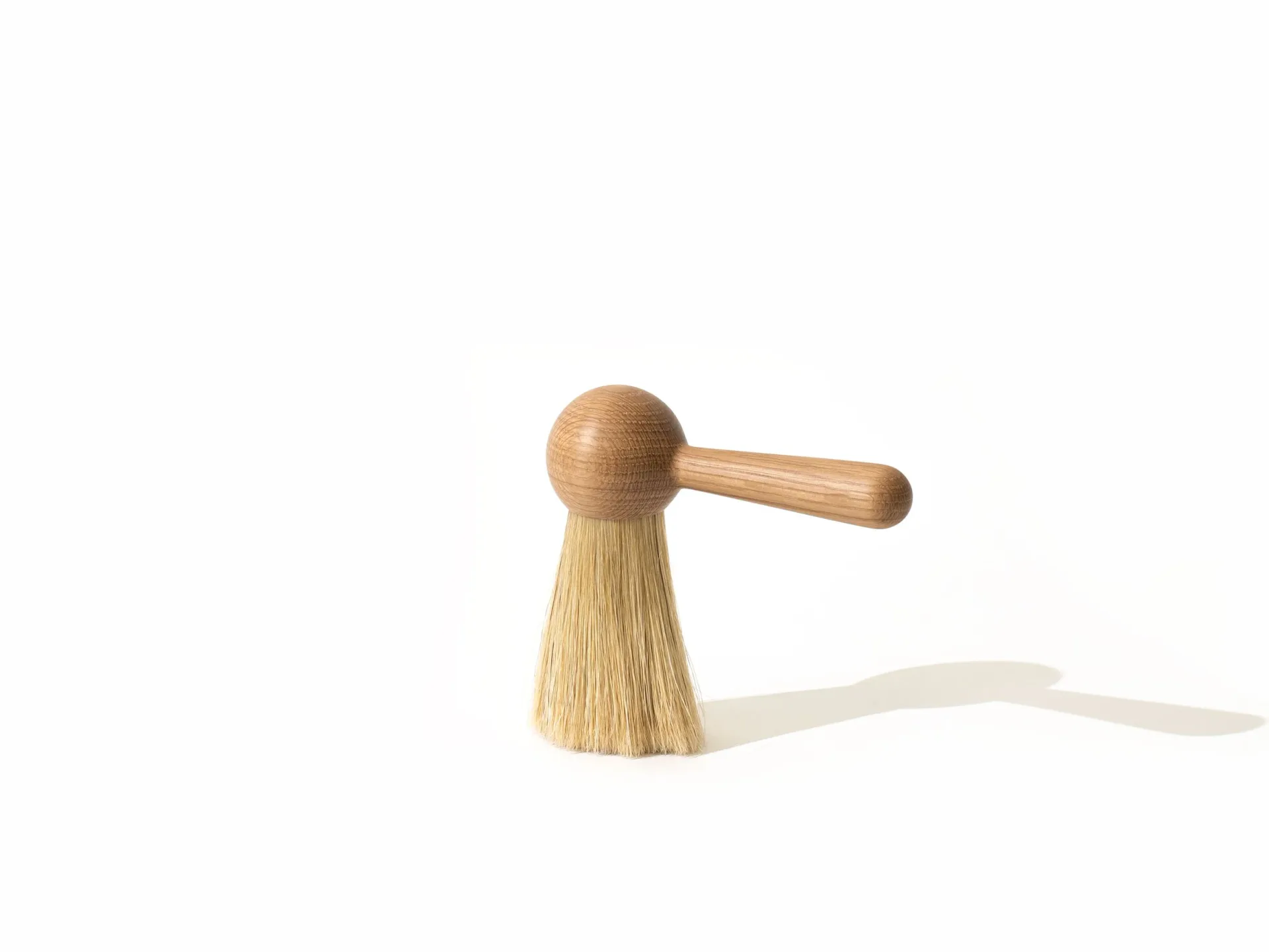 Wooden brush with spherical handle and natural bristles casting a soft shadow against white background, product design by Pat Kim.