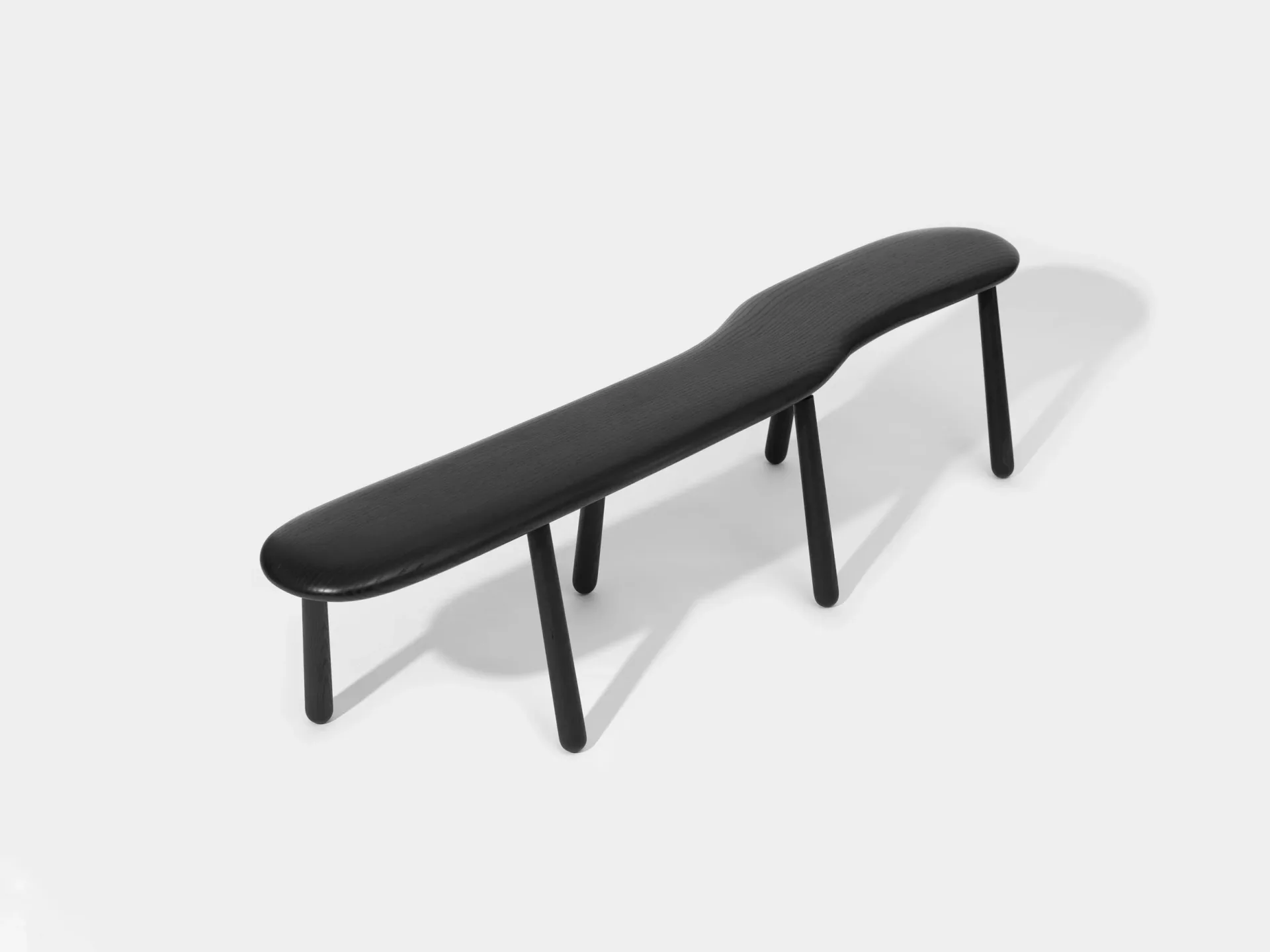 A long curved black bench with wooden legs cast against a light gray background, featuring synthetic shadow and smooth gradient surfaces characteristic of a 3D rendering by Pat Kim.