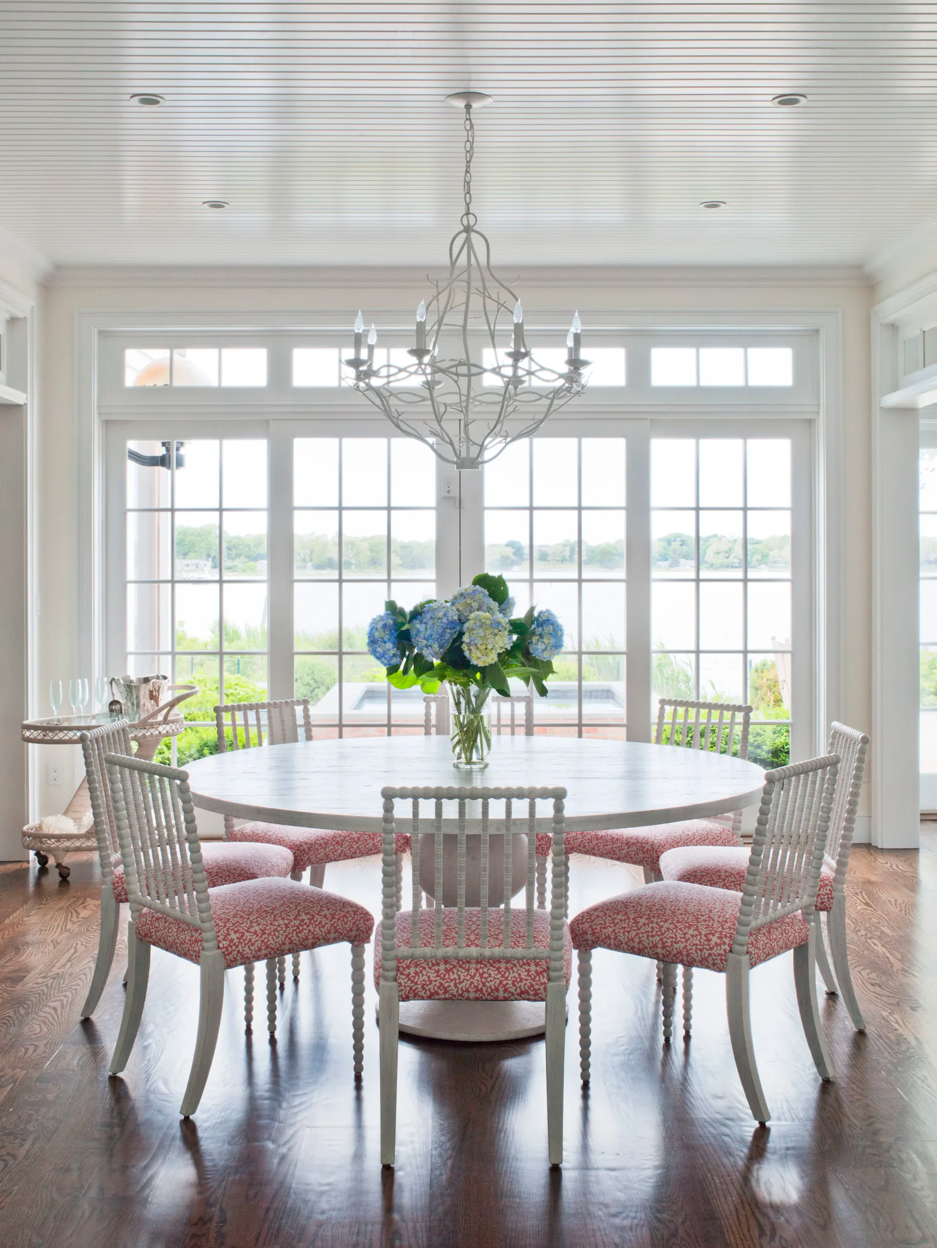 Bright dining room with round white table, gray chairs with pink cushions, crystal chandelier, and expansive multi-pane windows overlooking water by Patricia McGrath.