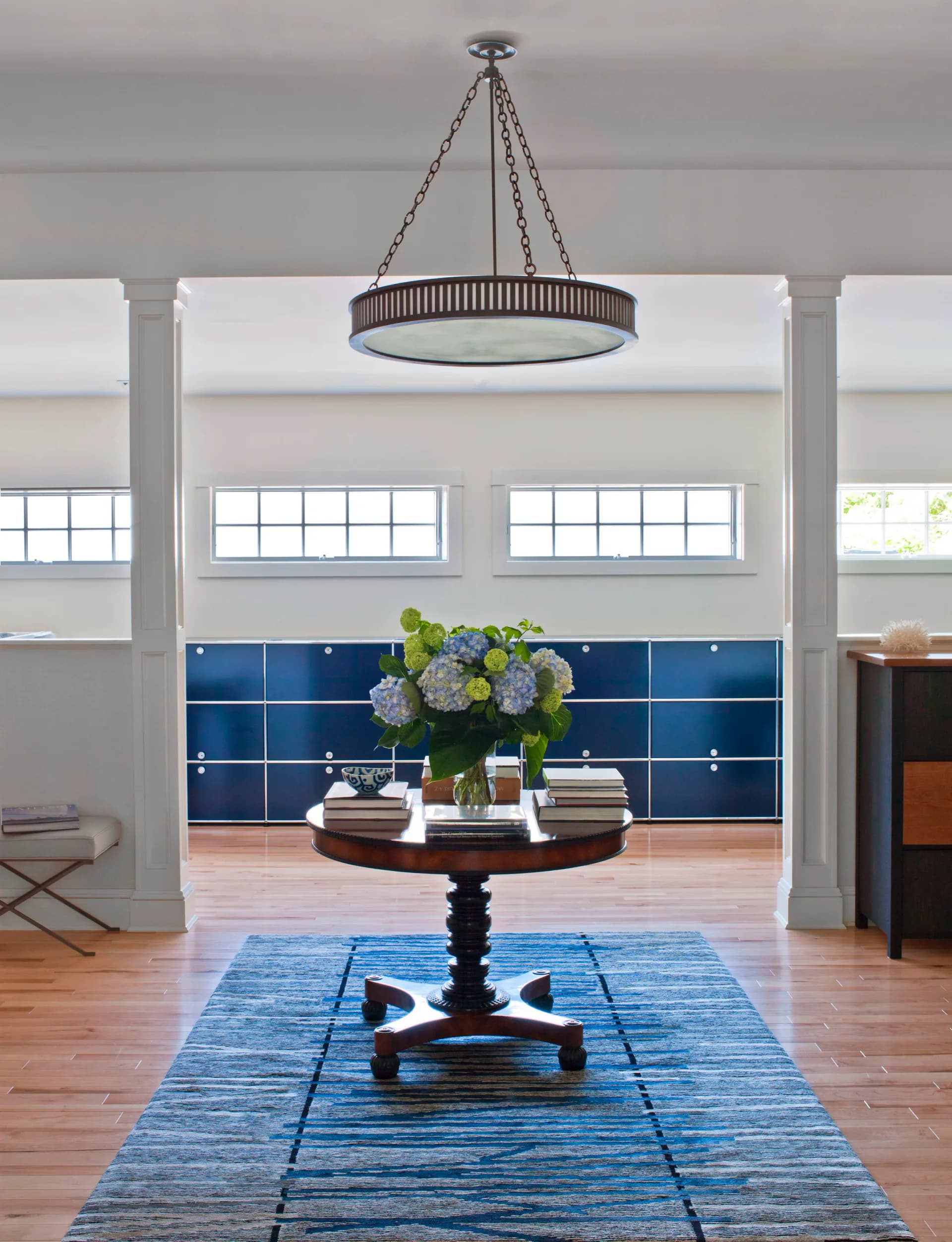 Modern foyer with pendant chandelier, blue tile backsplash, wooden pedestal table with hydrangeas, blue area rug, and tall columns by Patricia McGrath.