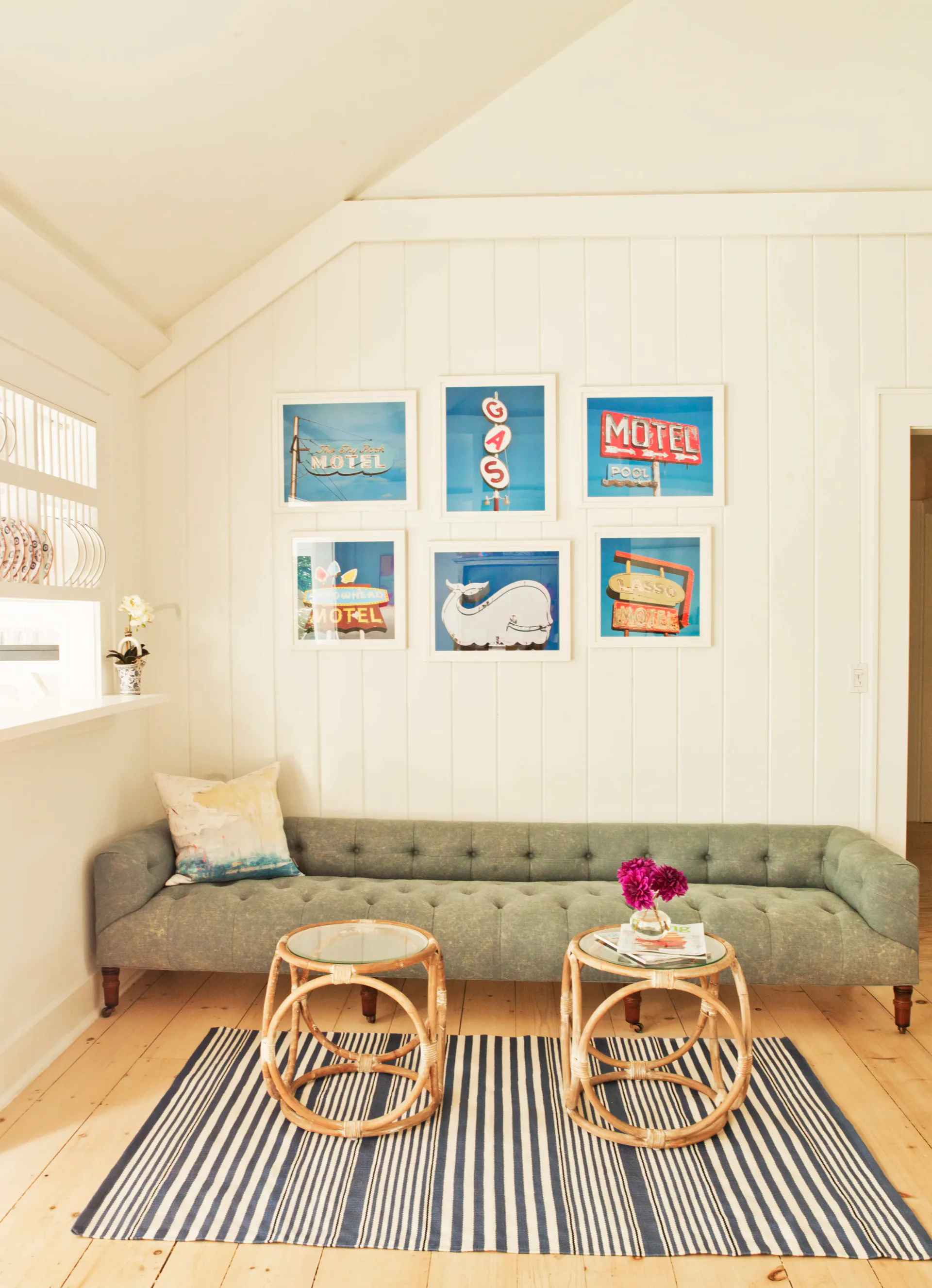 Light-filled living room with white shiplap walls, sage green tufted sofa, rattan side tables, striped rug, and gallery wall of vintage motel signage by Patricia McGrath.