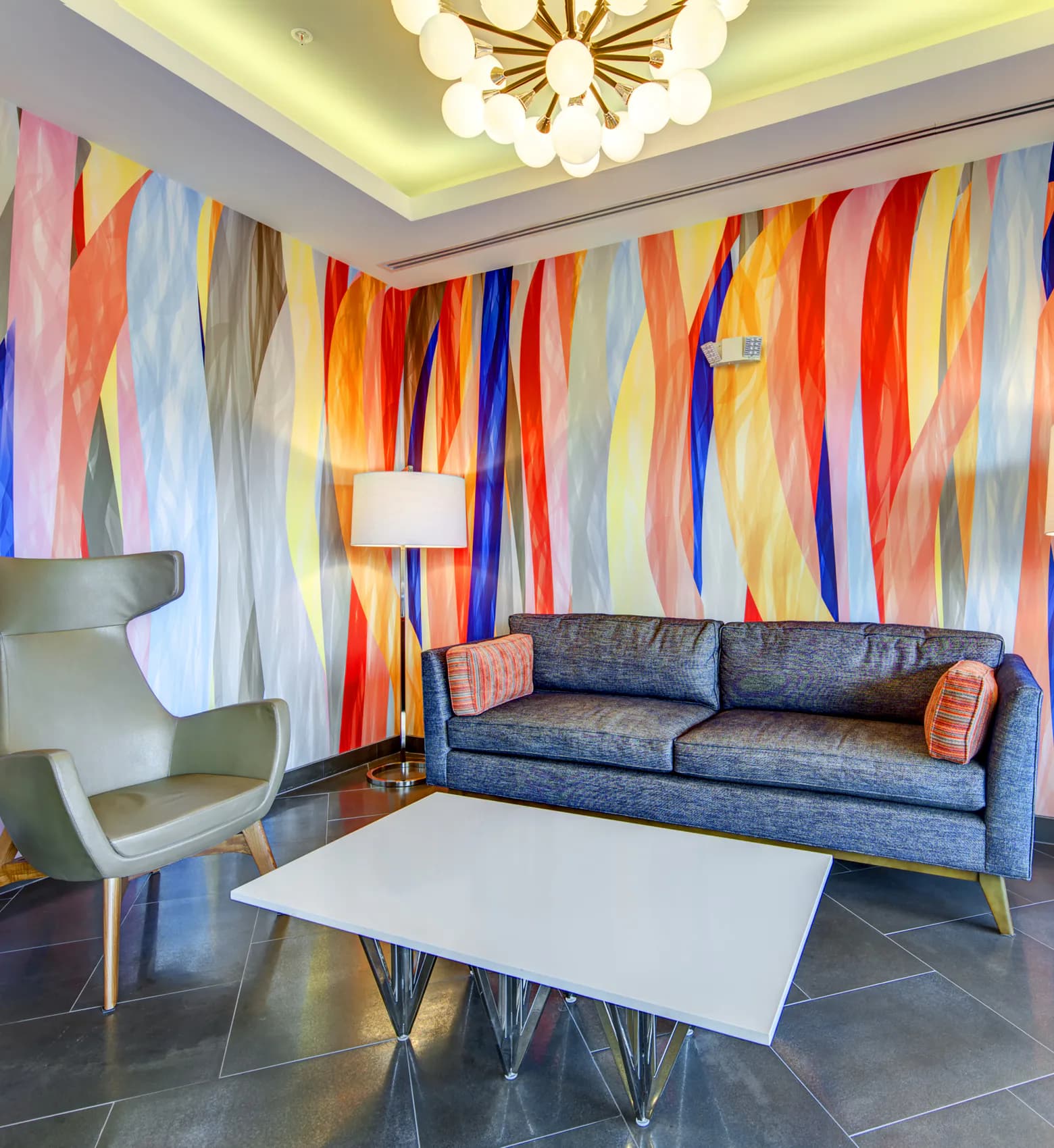 Modern living room with a blue upholstered sofa, white coffee table, sage green lounge chair, and vibrant multicolored striped accent wall installation by Patti Garner.