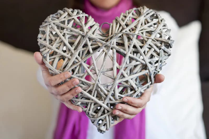 A woven white wicker heart-shaped decorative object held in hands, displaying intricate lattice craftsmanship by Patti Garner.