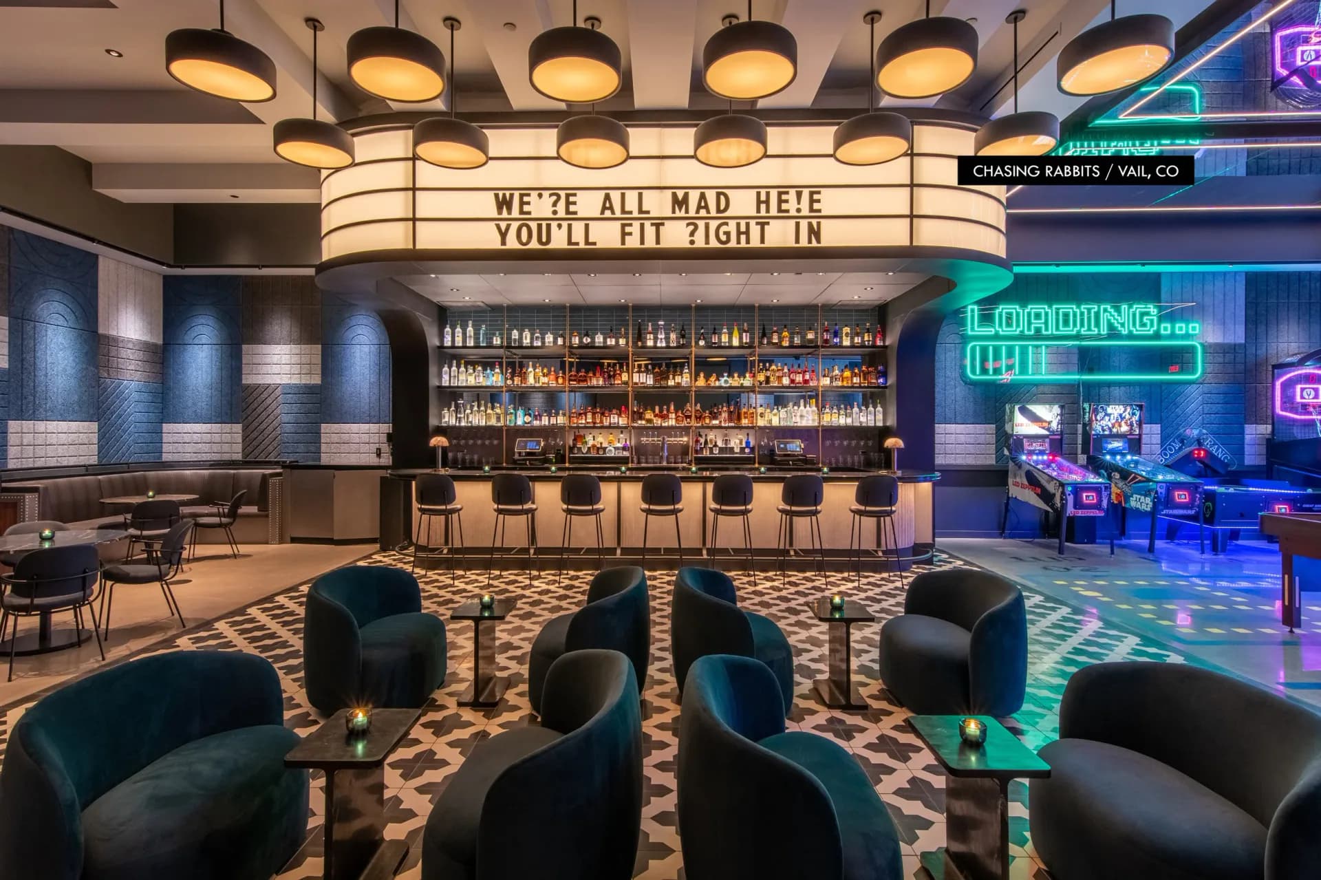 Modern bar lounge with dramatic lighting features a vintage marquee sign, curved black seating, patterned flooring, and neon accents, designed by Paul Gregory.