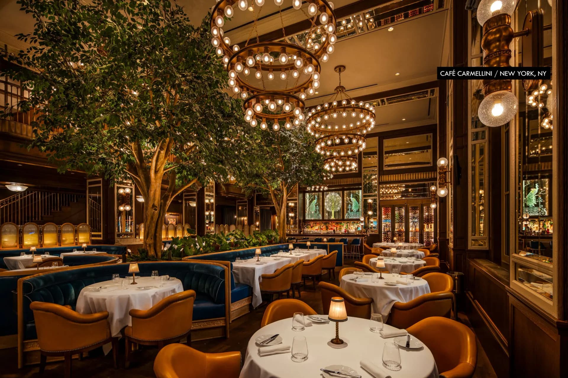 Elegant fine dining restaurant interior featuring warm brass tones, green foliage, sculptural pendant lighting, cognac leather banquettes, and white-draped tables by Paul Gregory.