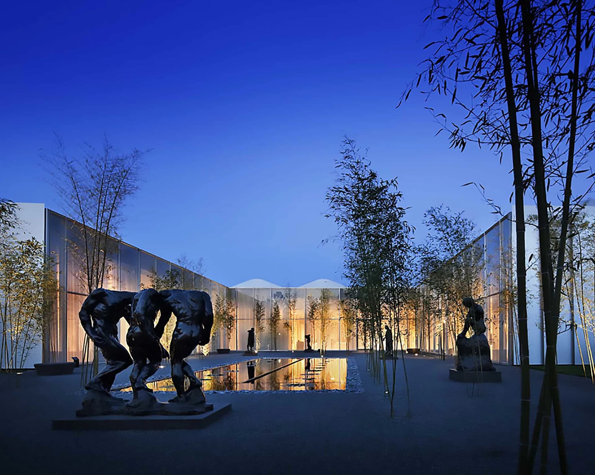 Modern architectural courtyard with illuminated stone walls, reflective water feature, sculptural bronze figures, and tall bamboo plantings at dusk by Paula Martinez-Nobles.