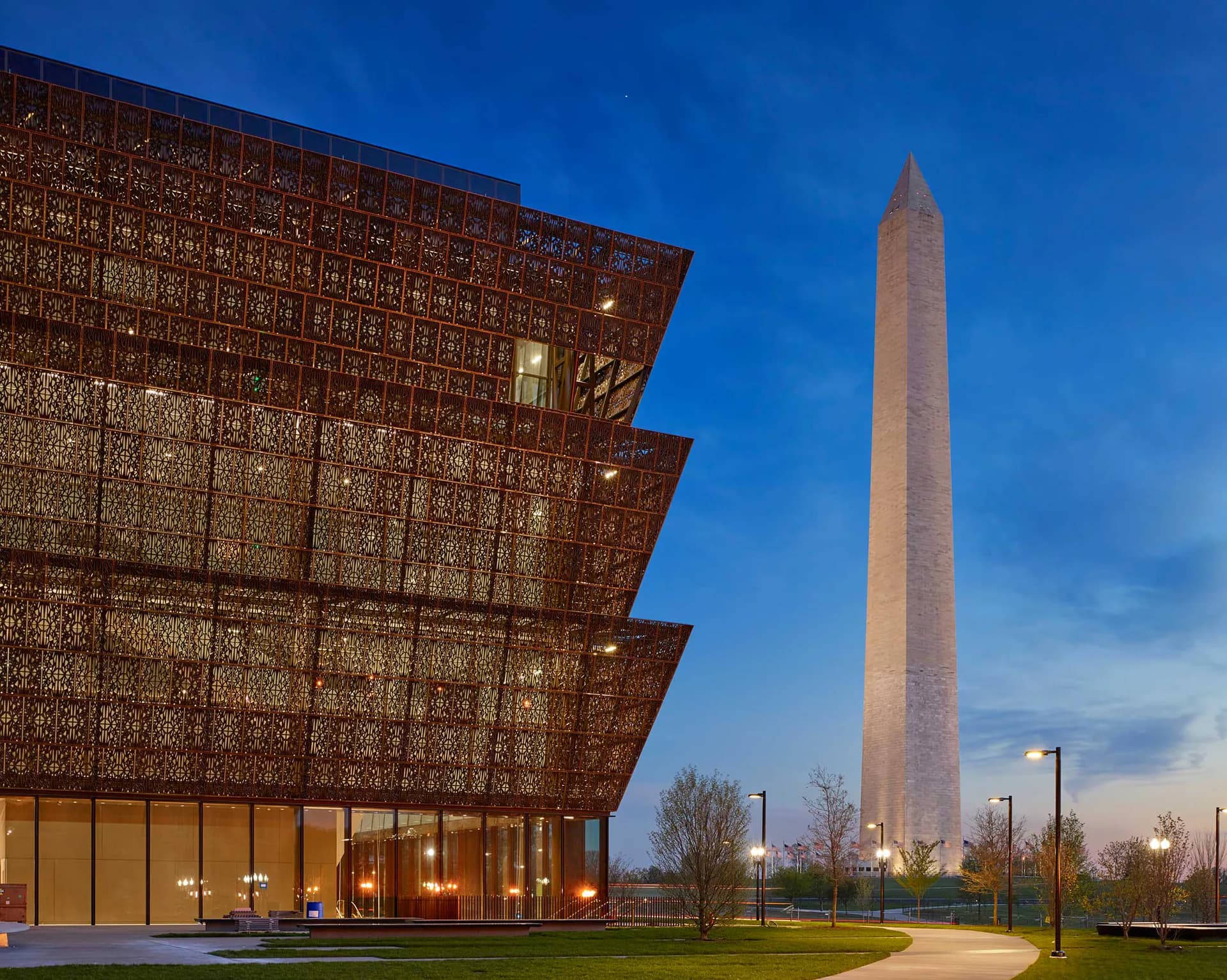 Modern brick museum building with geometric perforated facade beside Washington Monument at dusk, designed by Paula Martinez-Nobles.