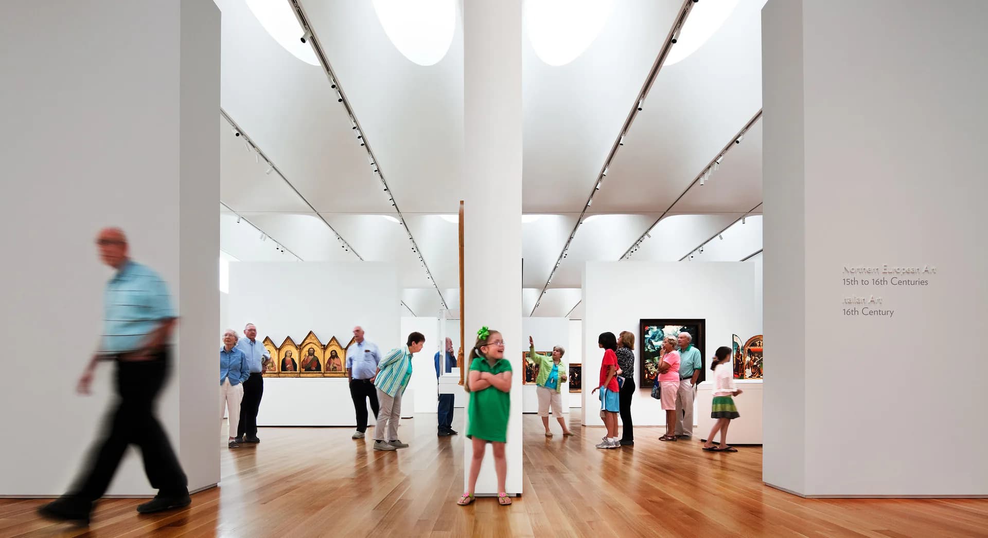 Bright, minimalist gallery space with soaring white ceilings, natural skylights, wooden floors, and display installations, filled with visitors by Paula Martinez-Nobles.