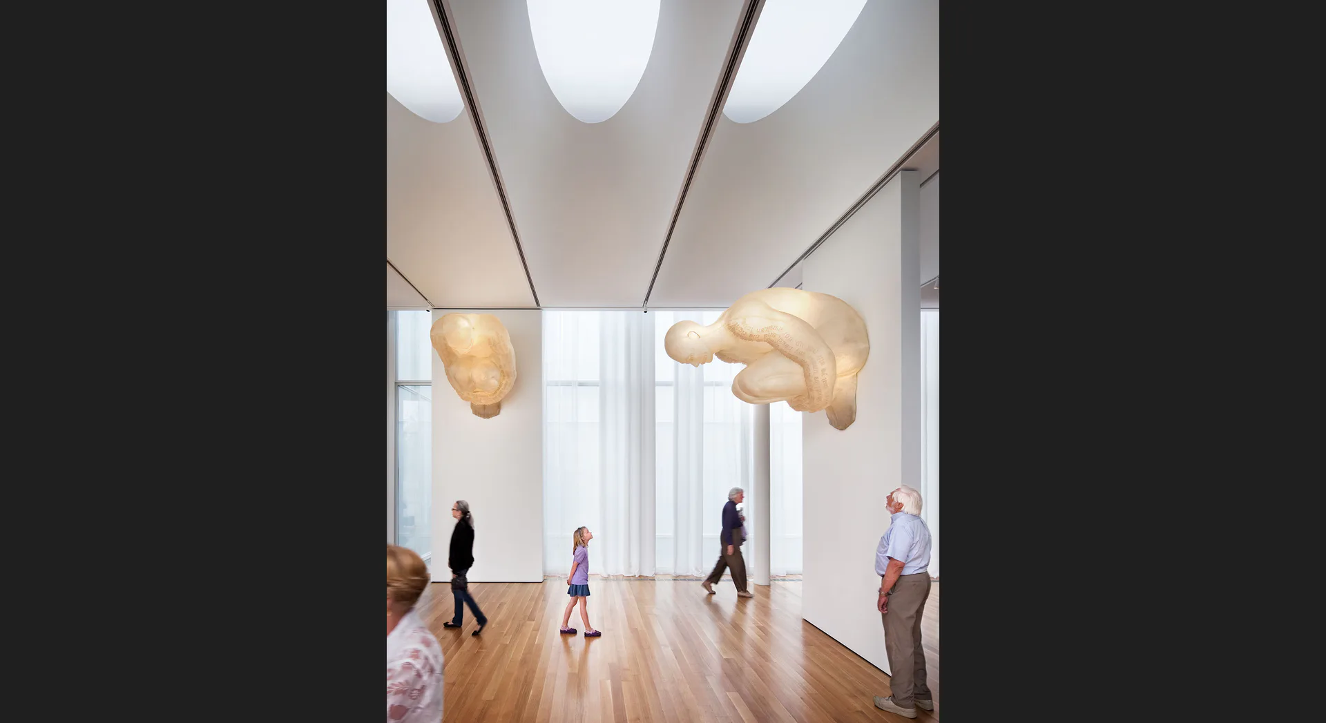 Minimalist gallery space with wooden floors, suspended organic sculptural forms in warm wood tones, and modern linear ceiling elements by Paula Martinez-Nobles.