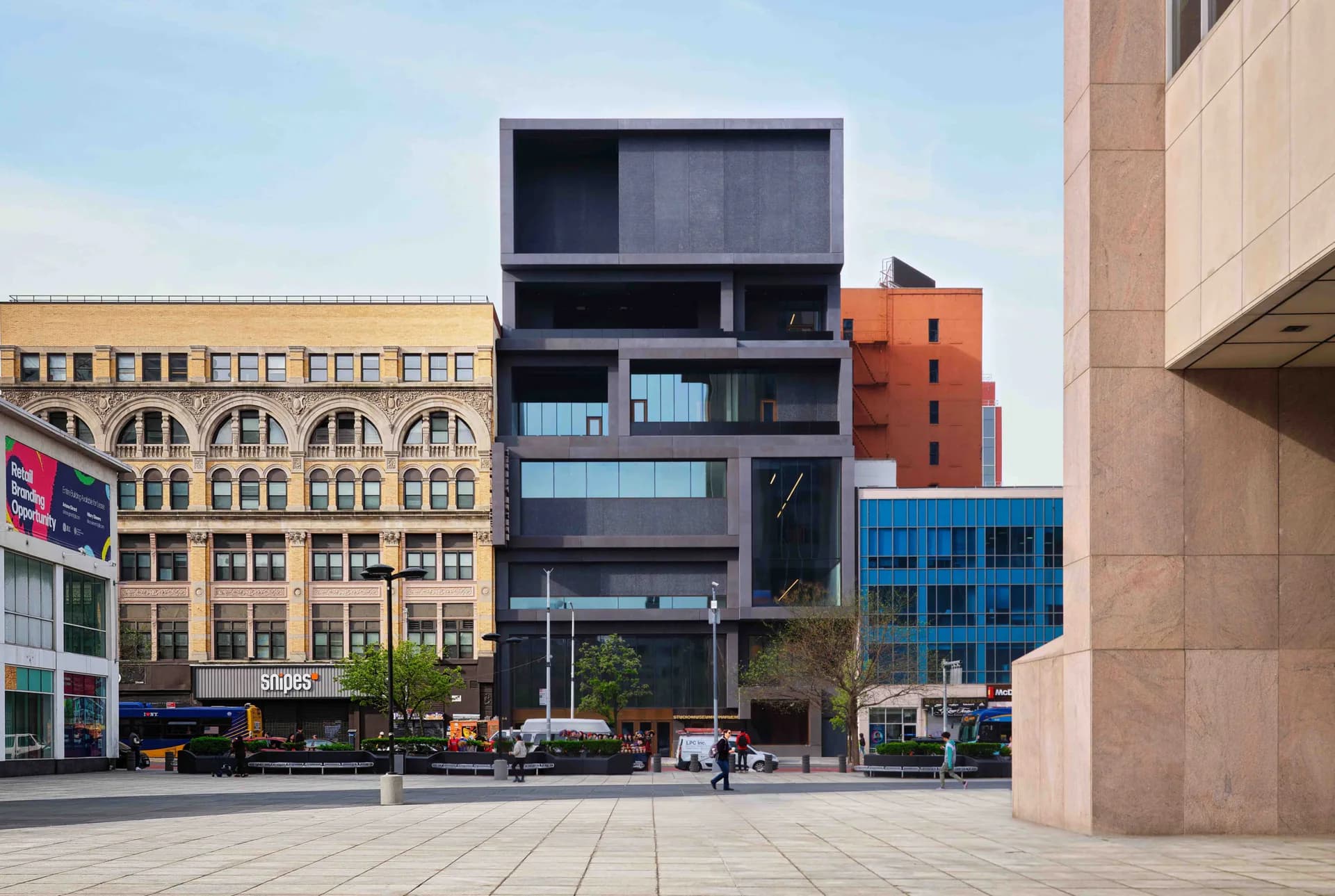 Modern black-clad building with horizontal window bands stands among colorful historic and contemporary architecture in an urban plaza, designed by Paula Sanchez.