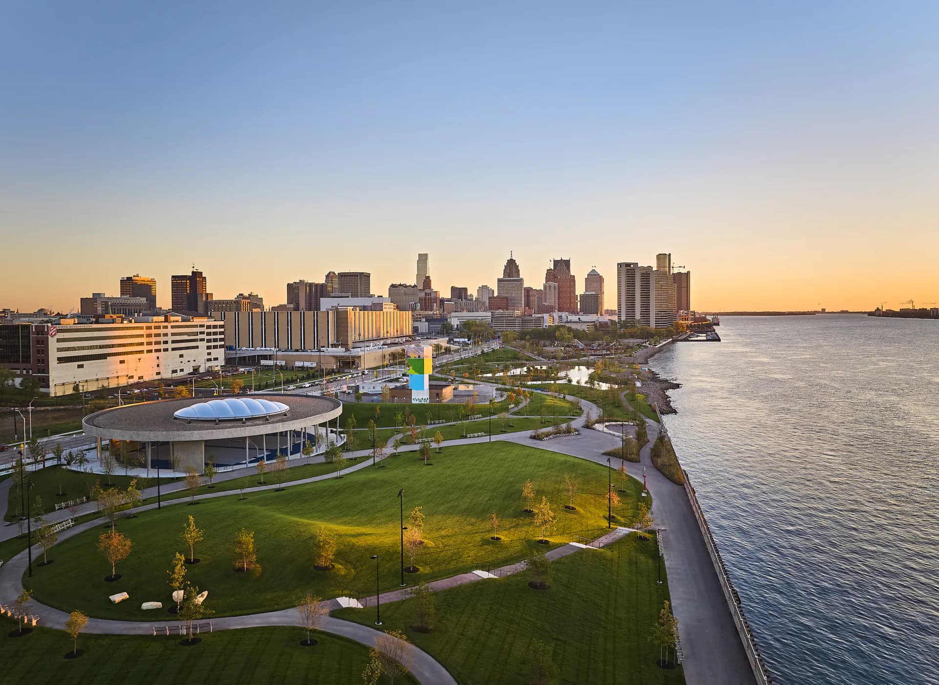 Waterfront park with illuminated landscape features, green lawn, and modern curved architecture overlooking downtown skyline at dusk by Paula Sanchez.