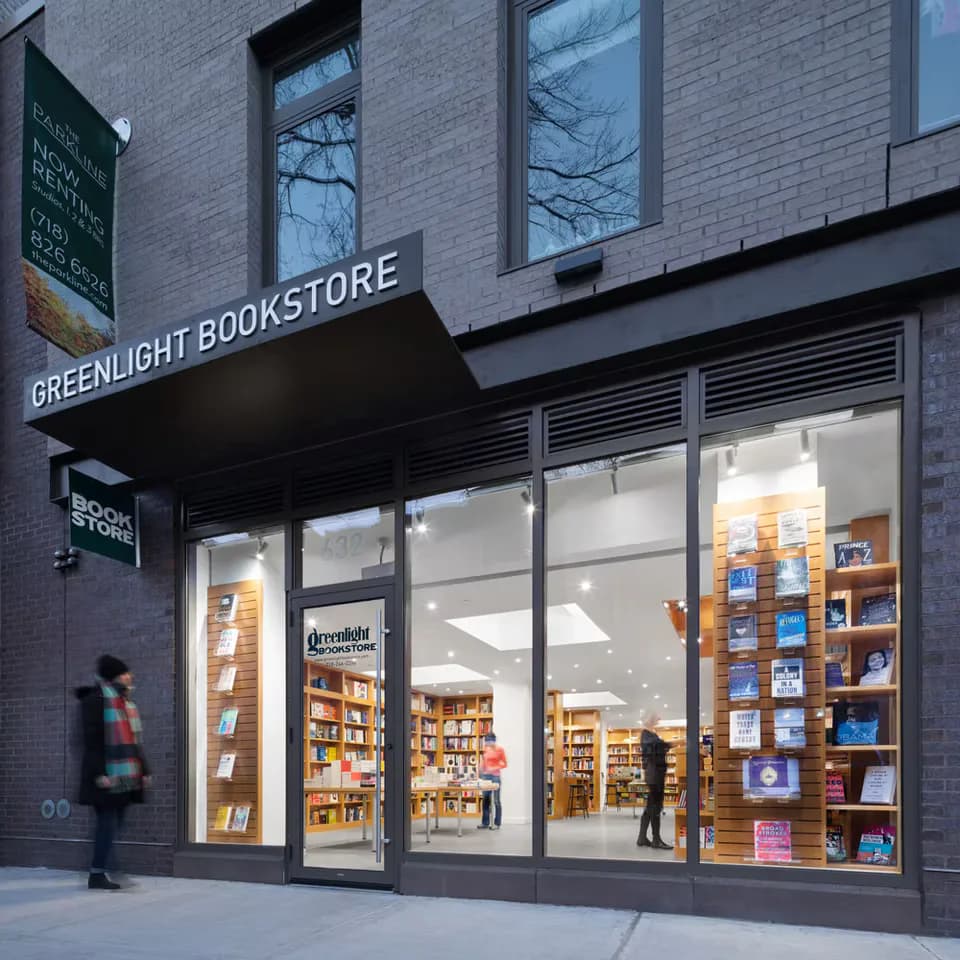 Greenlight Bookstore storefront with large windows, wooden shelving displays, warm interior lighting, and brick facade by Peiheng Tsai.