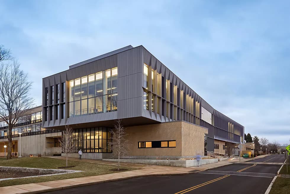 Modern institutional building with warm brick base and dark metal-clad upper floors featuring extensive glazing, illuminated interiors visible at dusk, by Peiheng Tsai.