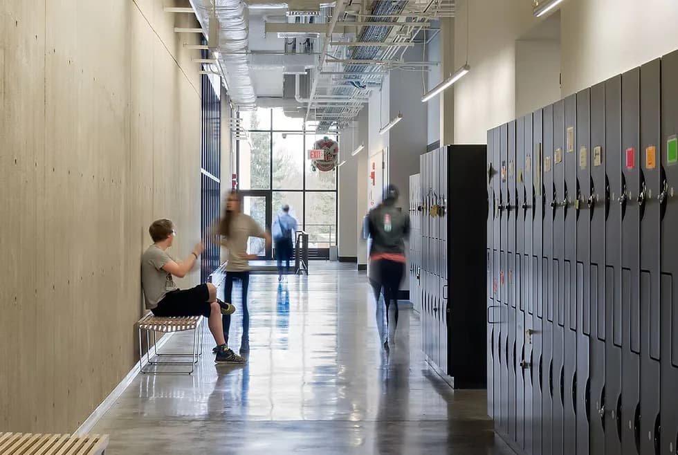 Modern school corridor with polished concrete floors, light wood and metal lockers, natural light from windows, and students in motion by Peiheng Tsai.