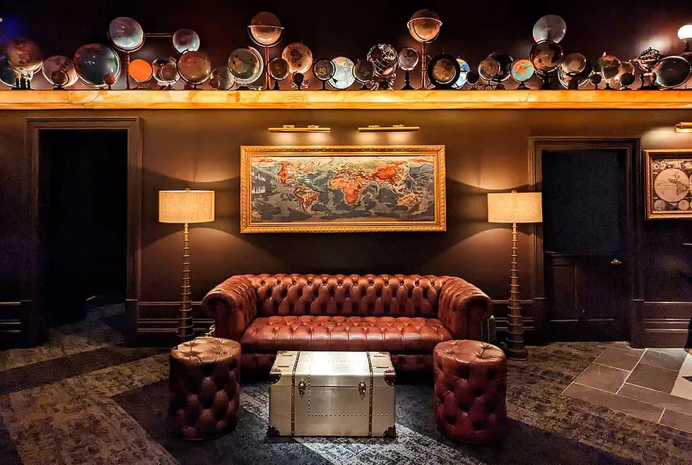 Luxury lounge featuring a cognac leather Chesterfield sofa, decorative plate wall display, brass floor lamps, and abstract artwork in warm earth tones by Peiheng Tsai.