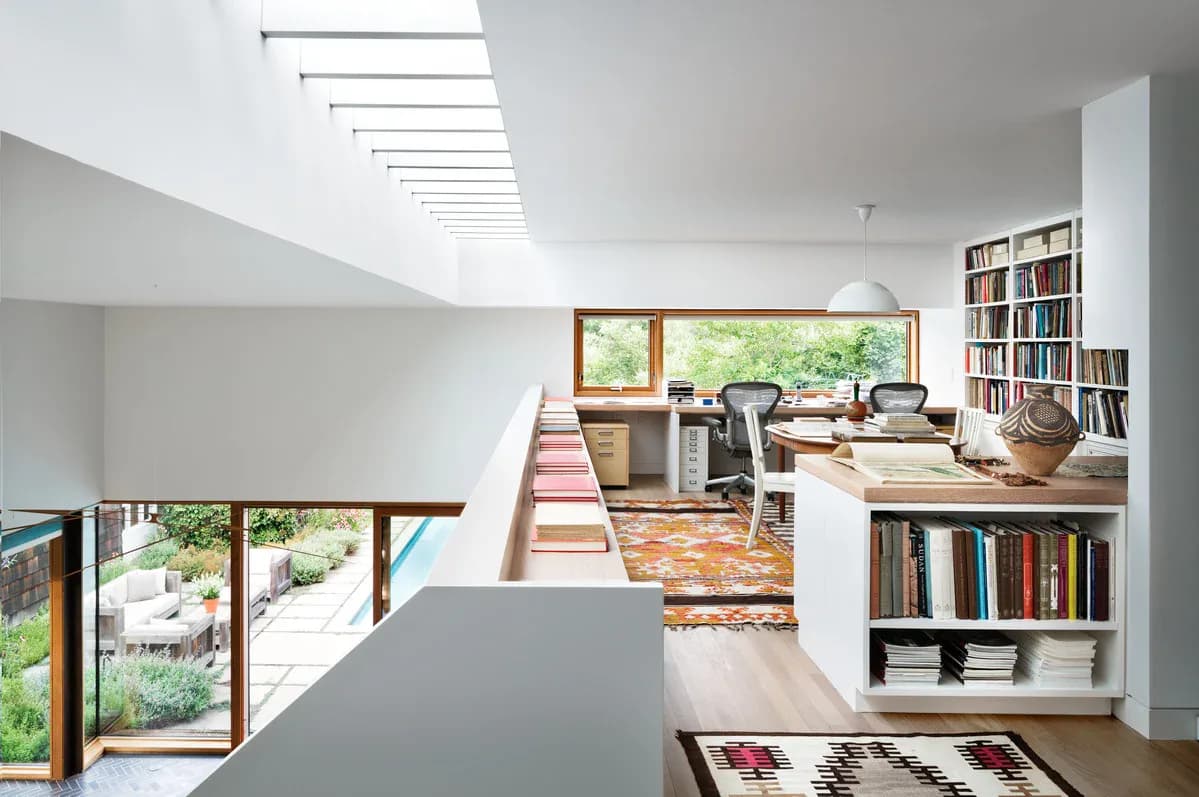 Bright multi-level loft interior with skylights, built-in bookshelves, wooden stairs, and large windows overlooking greenery by Perry Sayles.