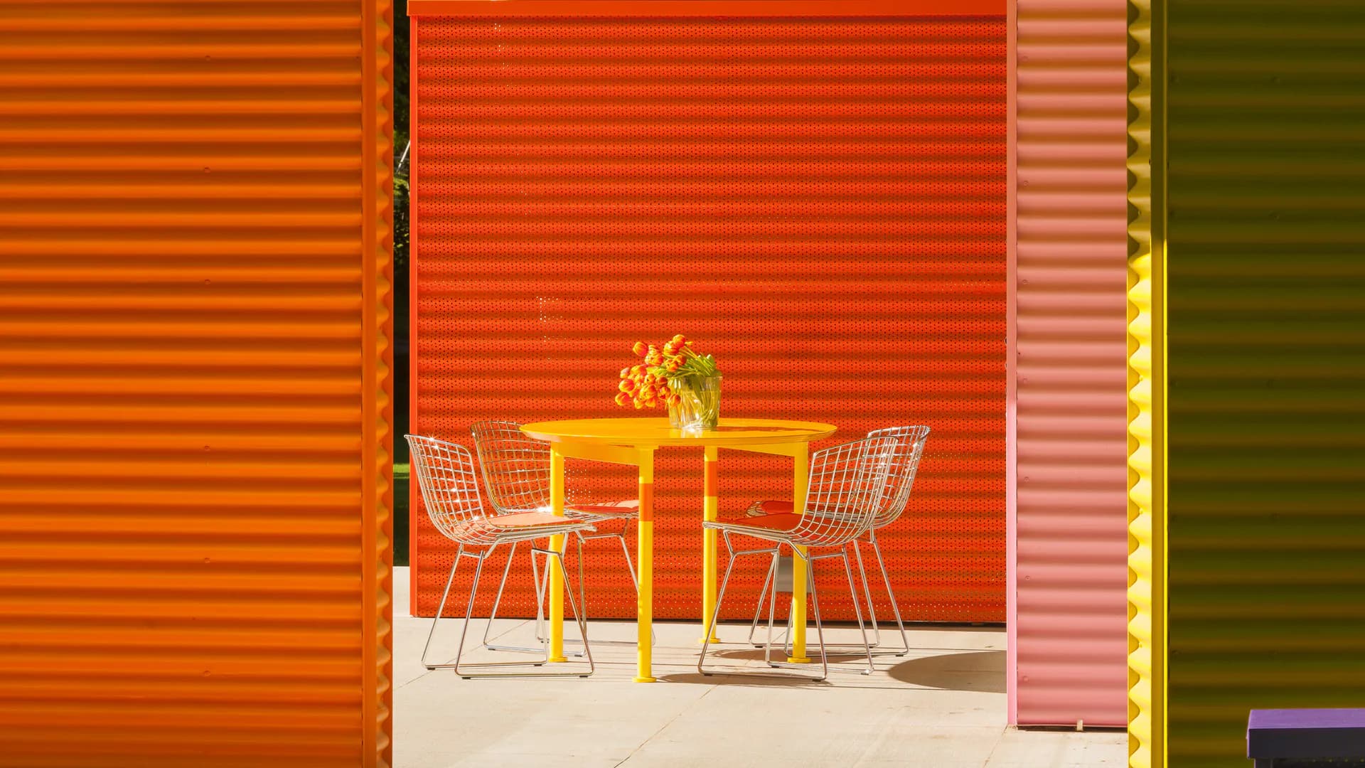 Minimalist interior with vibrant orange corrugated metal walls, yellow table, and patterned chairs in a bright, synthetic-looking space by Peter Stamberg.