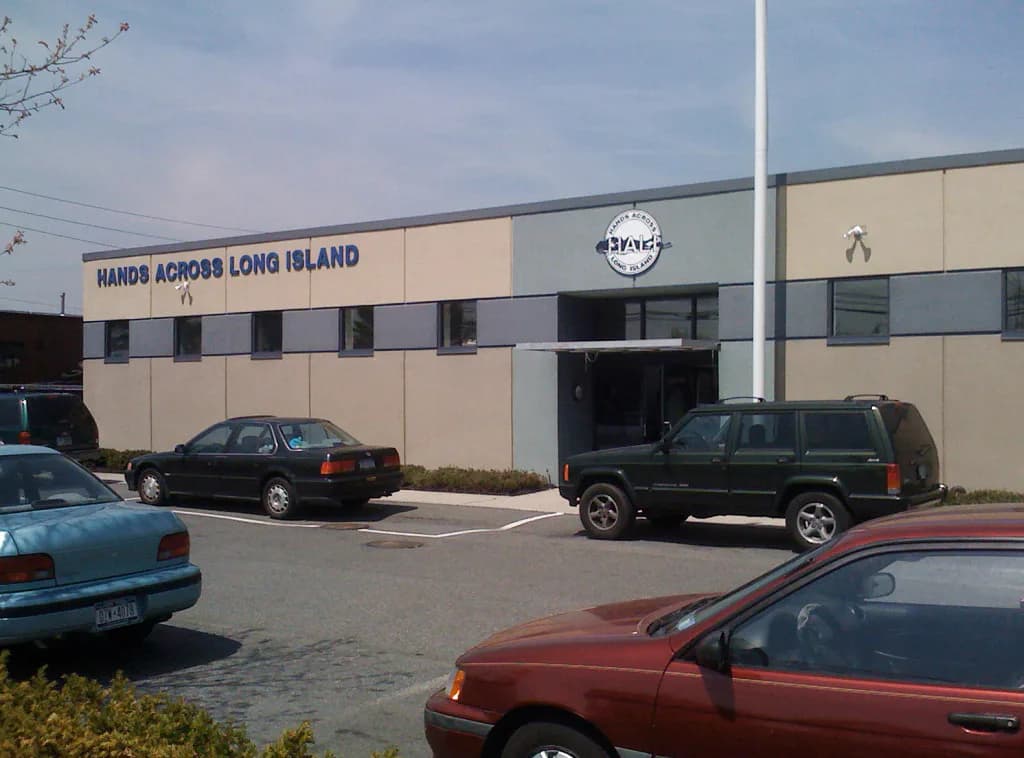 Exterior view of Hands Across Long Island commercial building with tan and gray facade, large windows, and parking lot with vehicles by Philip A. Mitropoulos.