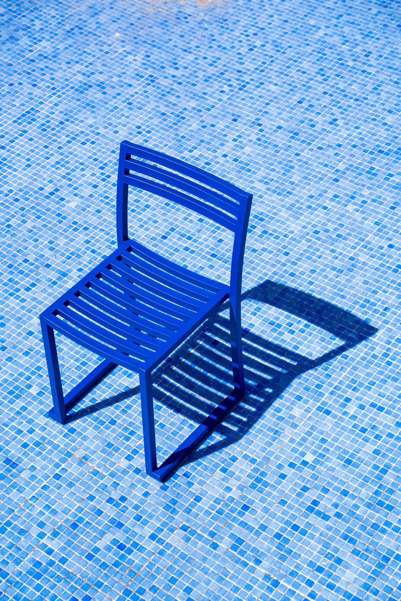 Bright blue slatted chair casting sharp shadow on mosaic pool tiles by Philippe Malouin.