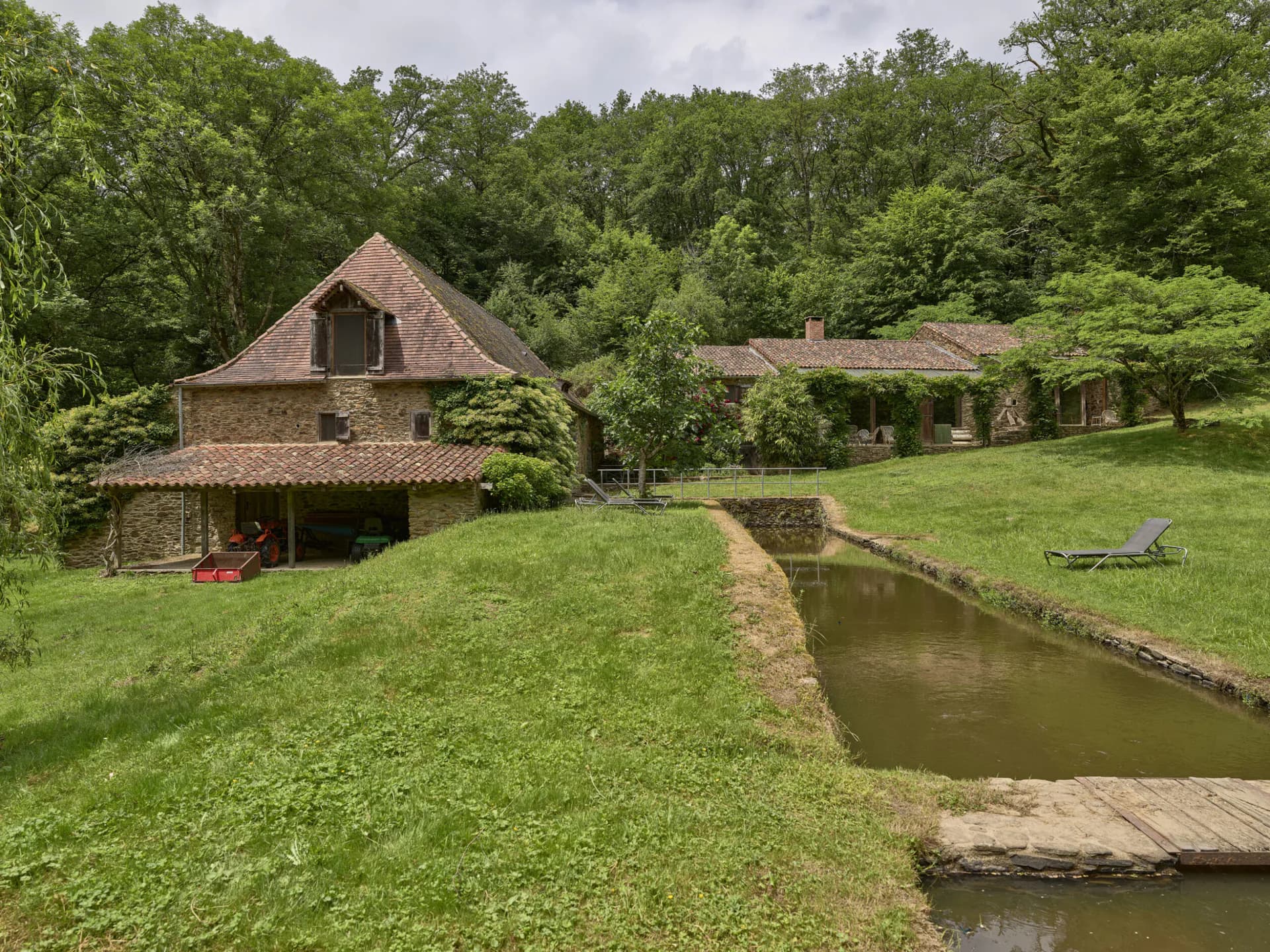 Rustic stone barn and farm buildings with terracotta tile roofs nestled in lush green countryside beside a pond, designed by Piet Hein Eek.