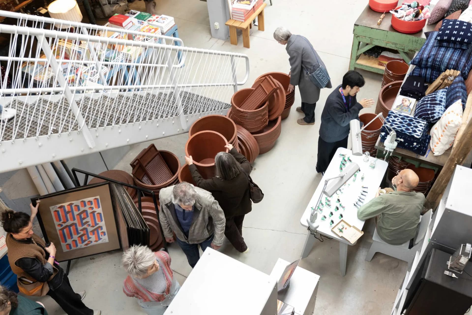 Overhead view of an exhibition or installation space with terracotta ceramic vessels and furniture pieces displayed on white flooring, visitors browsing the collection by Piet Hein Eek.
