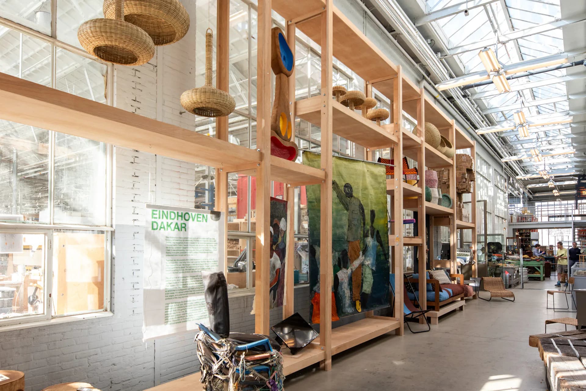 A bright, naturally-lit warehouse showroom with timber shelving units displaying furniture and home goods, featuring woven pendant lights and large windows, designed by Piet Hein Eek.