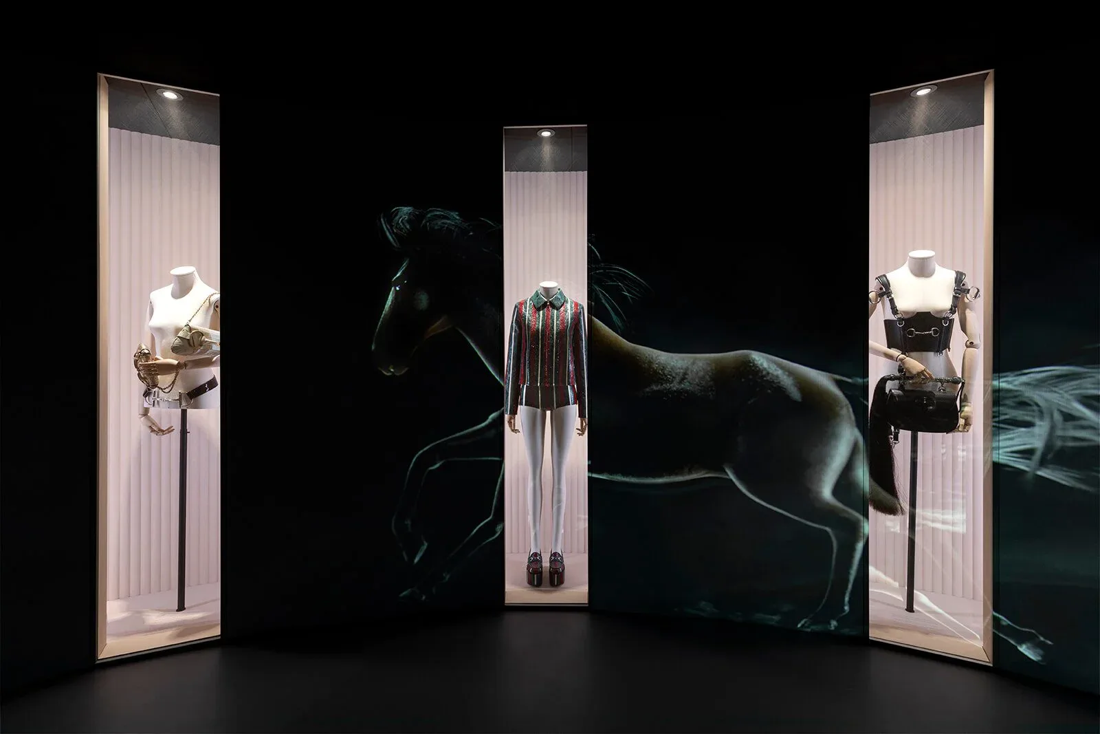 Dark dramatic exhibition installation featuring illuminated vertical display cases with sculptural objects and bronze horse statues by Polina Zakh.