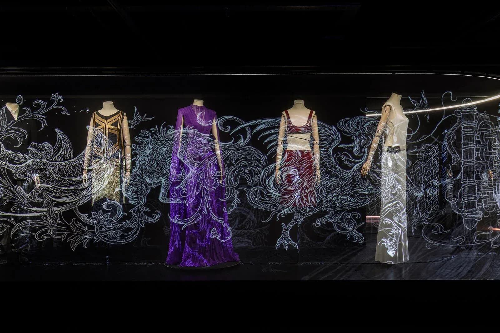 Exhibition display of five elaborate gowns on mannequins against an ornate black backdrop with intricate silver line drawings, featuring gold, purple, and embellished fabrics by Polina Zakh.