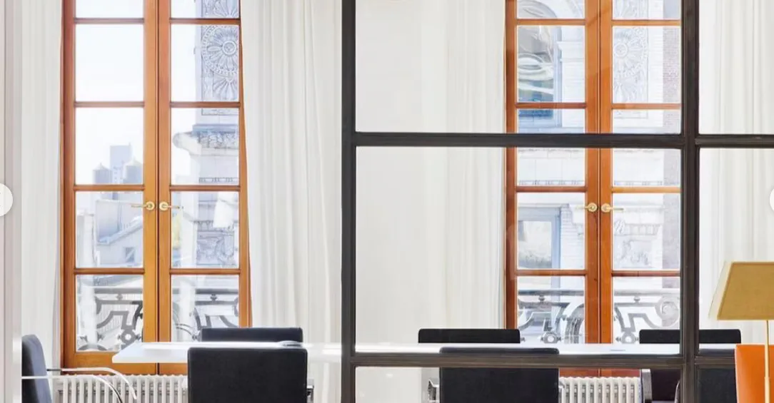 Modern office interior featuring floor-to-ceiling windows with wood frames, black desk workstations, and minimalist furnishings overlooking a city skyline by Prosha Korangy.