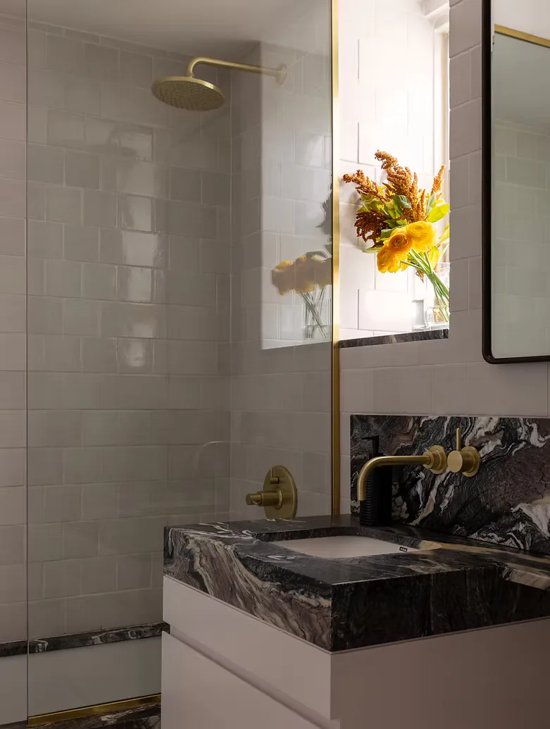 Modern bathroom with marble vessel sink, white cabinetry, subway tile shower, brass fixtures, and fresh flowers in natural light by Prosha Korangy.