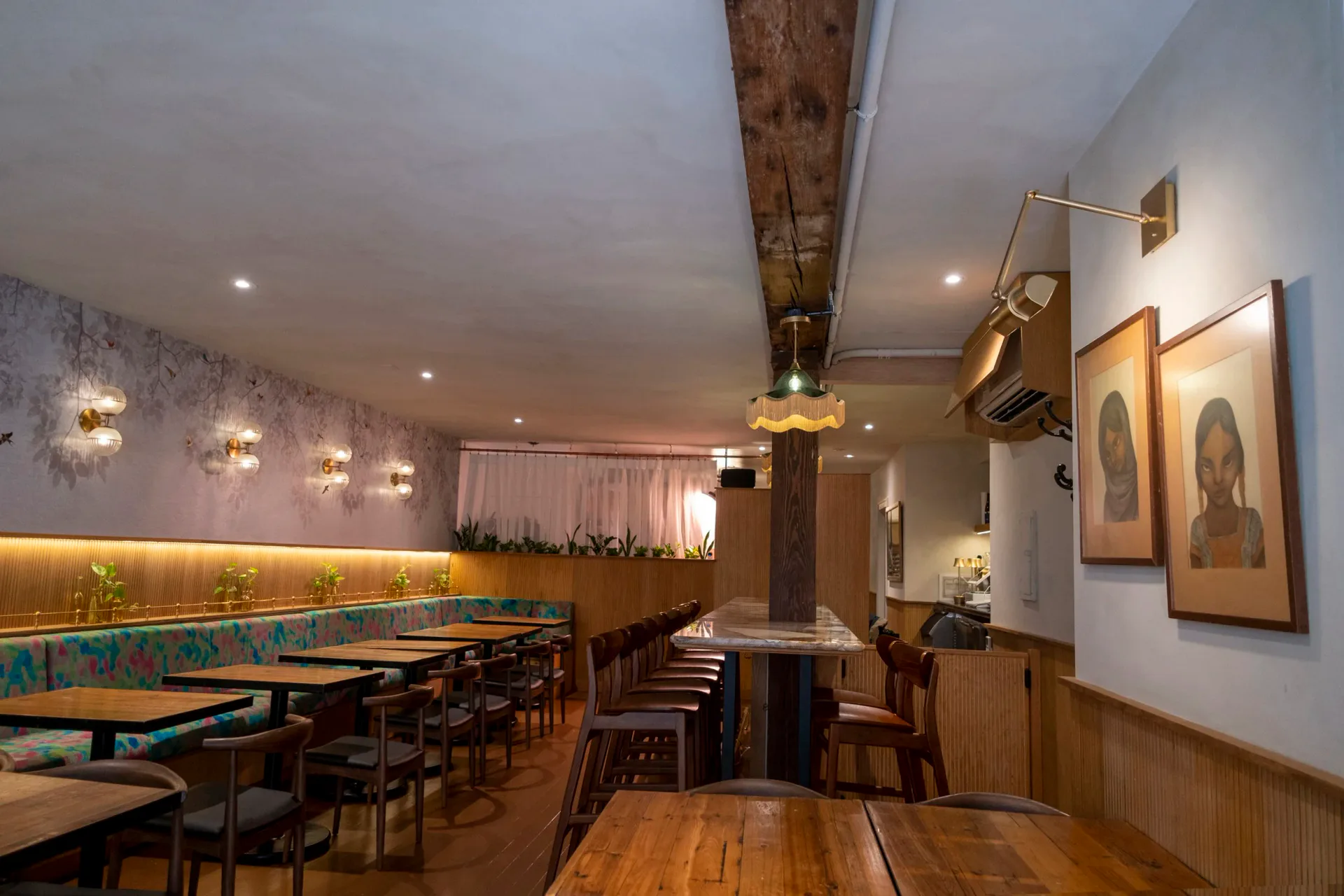 Modern restaurant interior with warm wood elements, brass lighting fixtures, teal banquettes, and framed artwork on white walls by Prutha Raithatha.