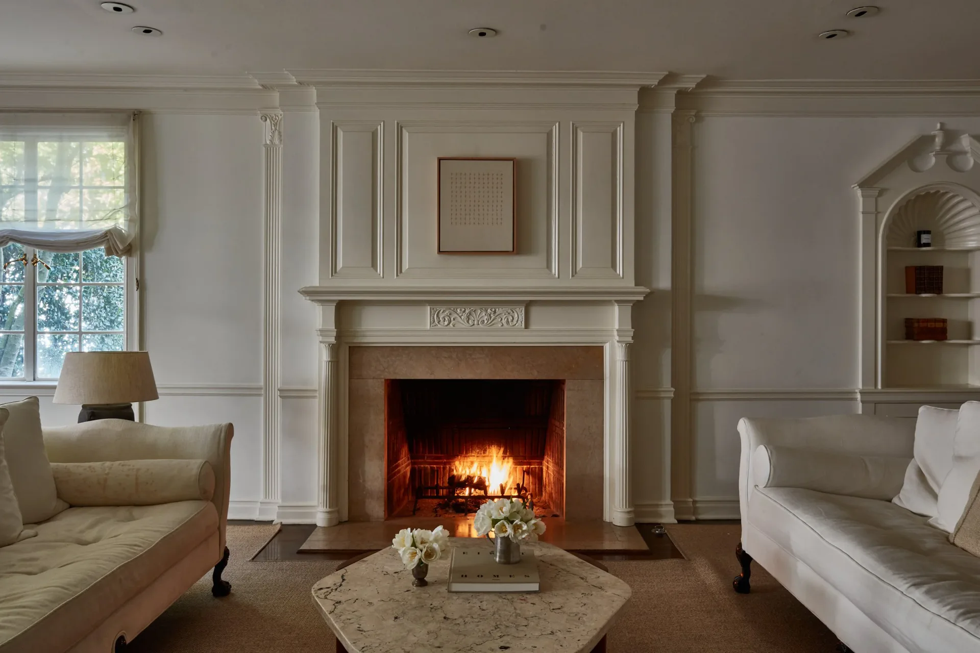 Elegant living room with cream paneled walls, marble fireplace with active fire, symmetrical seating, and arched alcove by Puja Clarke.