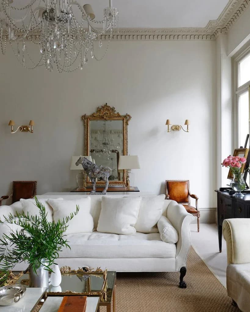 Elegant living room with white upholstered sofa, gilt-framed mirror above console table, crystal chandelier, brass wall sconces, and classical architectural details by Puja Clarke.