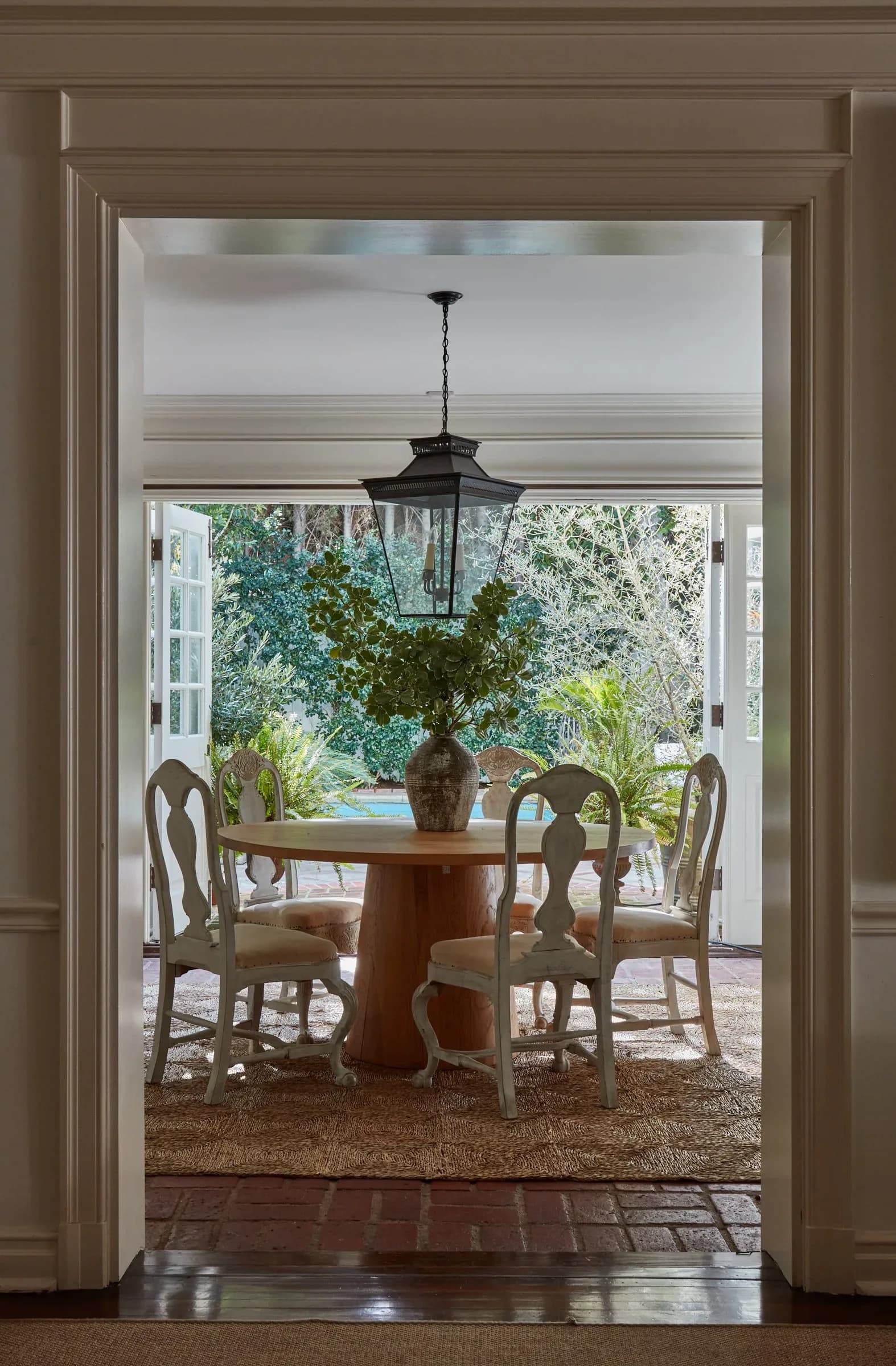 Elegant dining room with round wood table, period chairs, and pendant light opening to a poolside loggia with lush greenery by Puja Clarke.