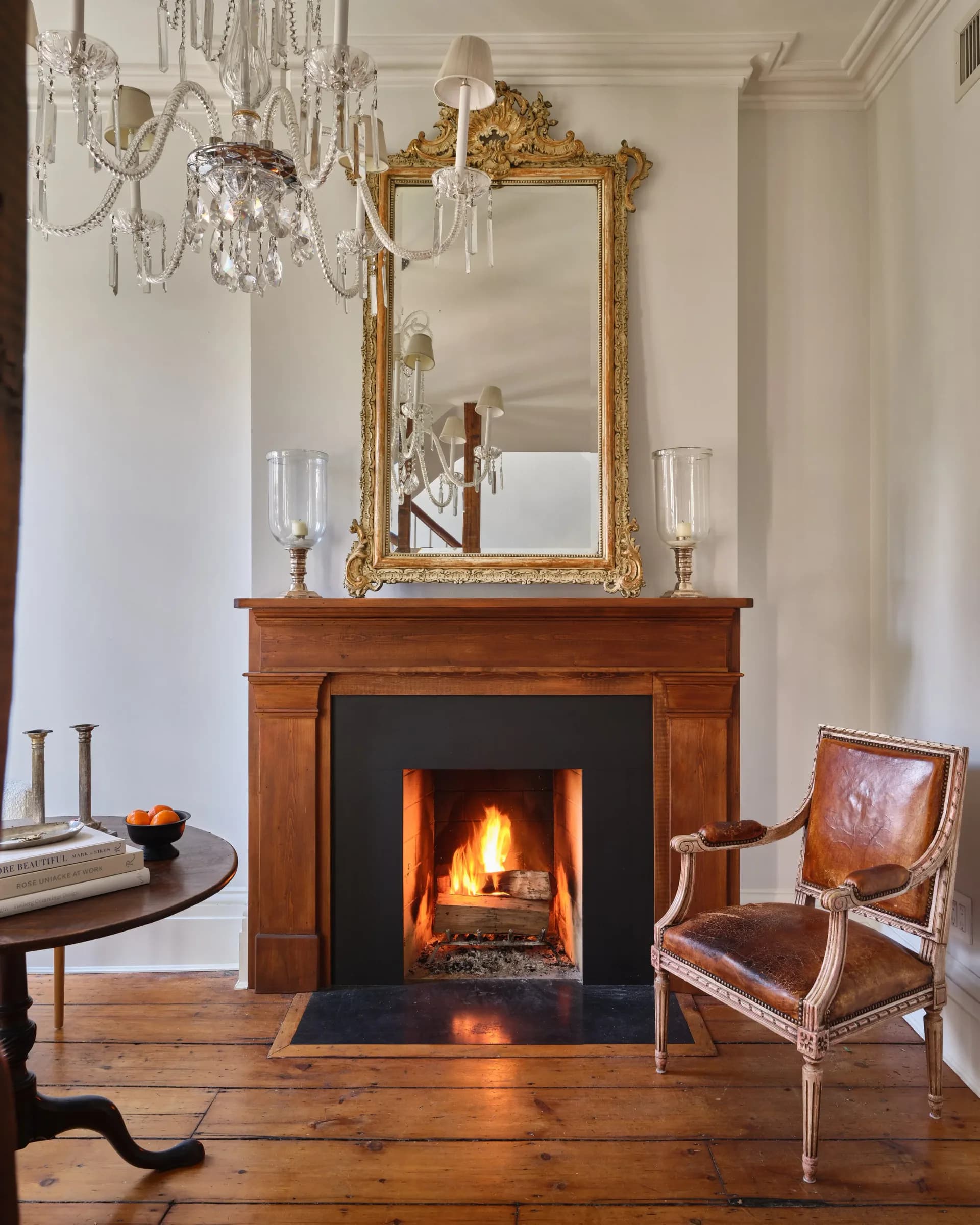 Elegant fireplace interior with warm wood mantel, ornate gold mirror, crystal chandelier, leather wingback chair, and glowing fire, designed by Puja Clarke.