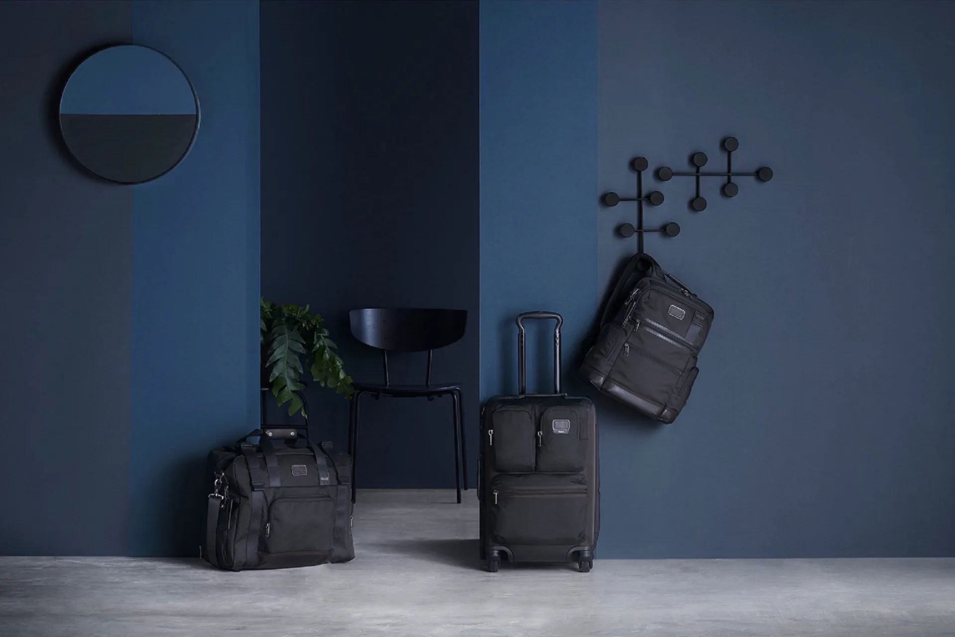 Modern interior with blue-gray walls displaying black luggage and travel bags alongside geometric wall sculpture and minimalist furniture by Quin Nichols.