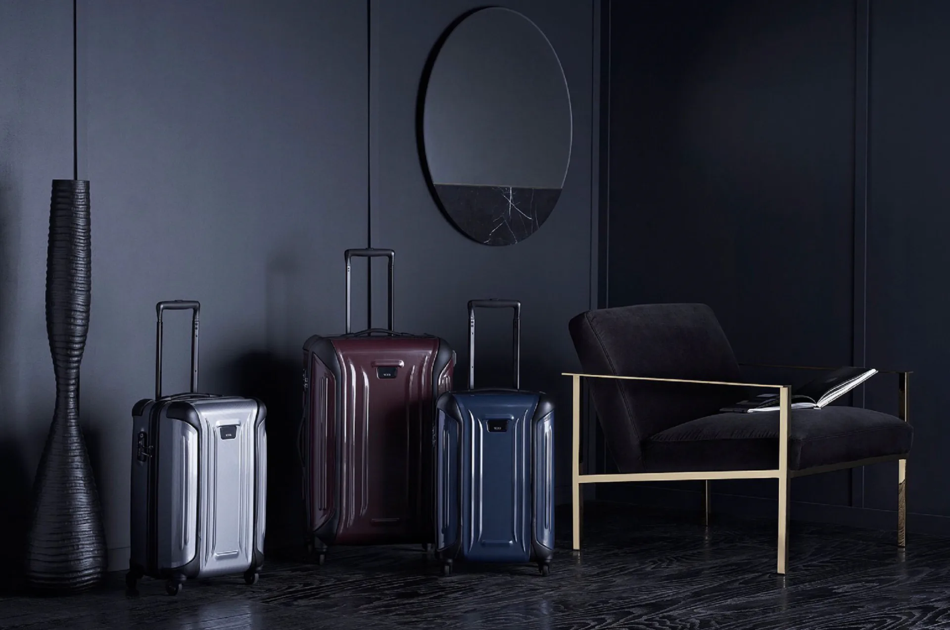 Moody dark interior with three luggage pieces, woven vase, oval mirror, and wooden chair with gold accents against charcoal walls by Quin Nichols.