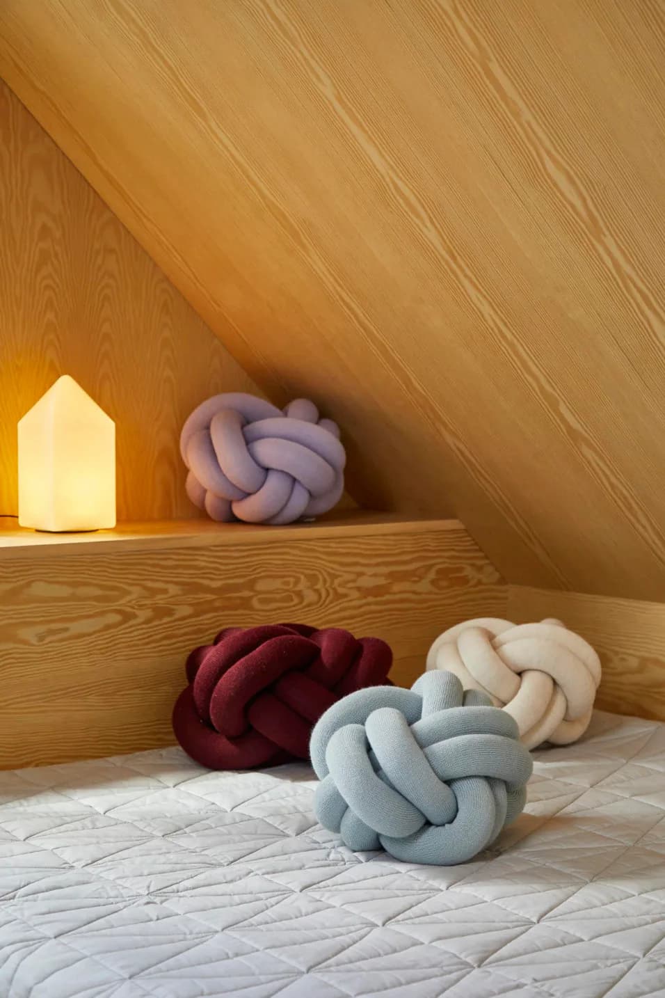 Cozy attic bedroom nook with sloped wooden ceiling, glowing house-shaped lamp, and colorful knotted throw pillows on quilted bedding by Quin Nichols.