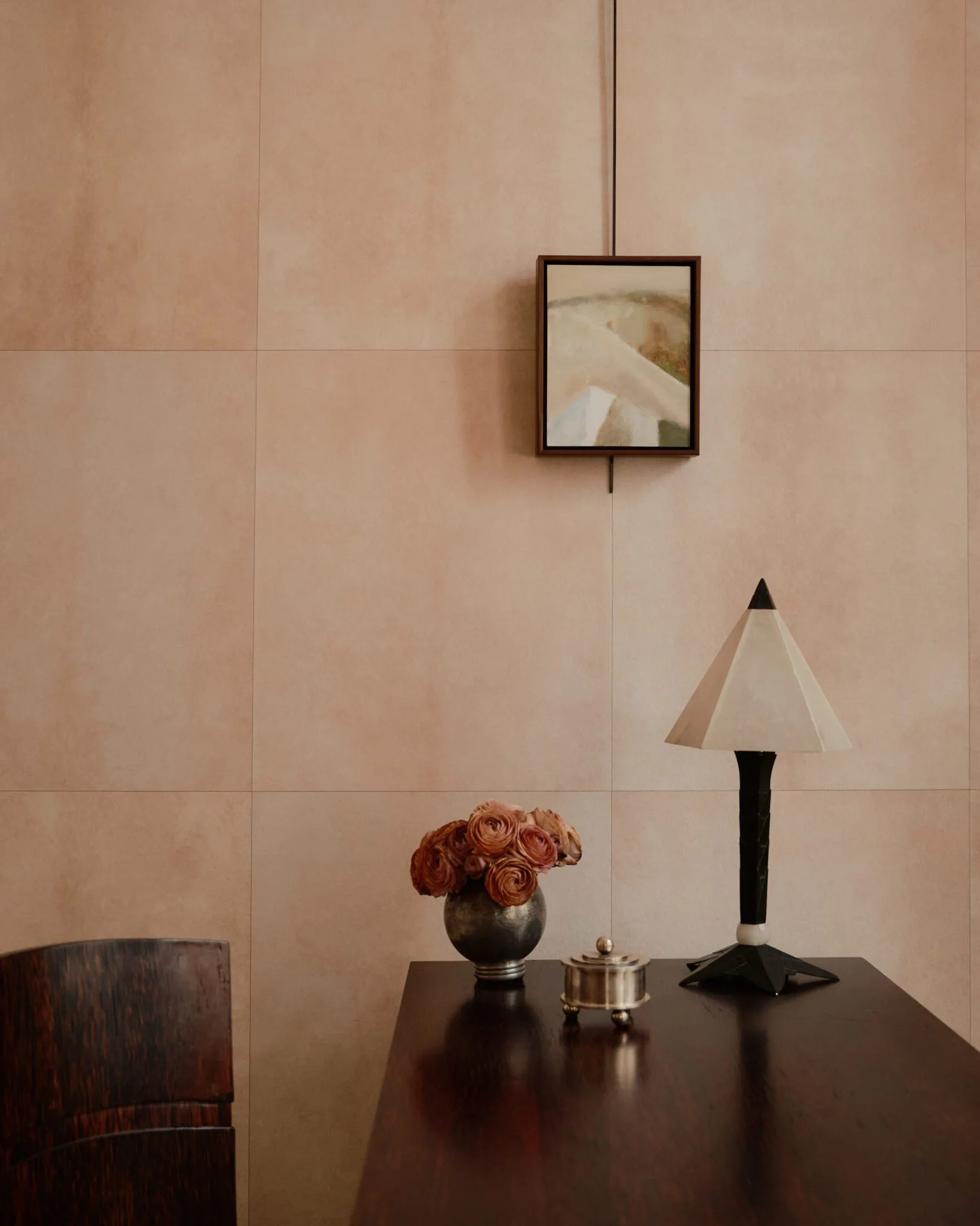 Minimalist interior with beige tiled wall, dark table displaying copper flowers in sphere vase, table lamp with cream shade, and framed artwork above by Rachel Cope.