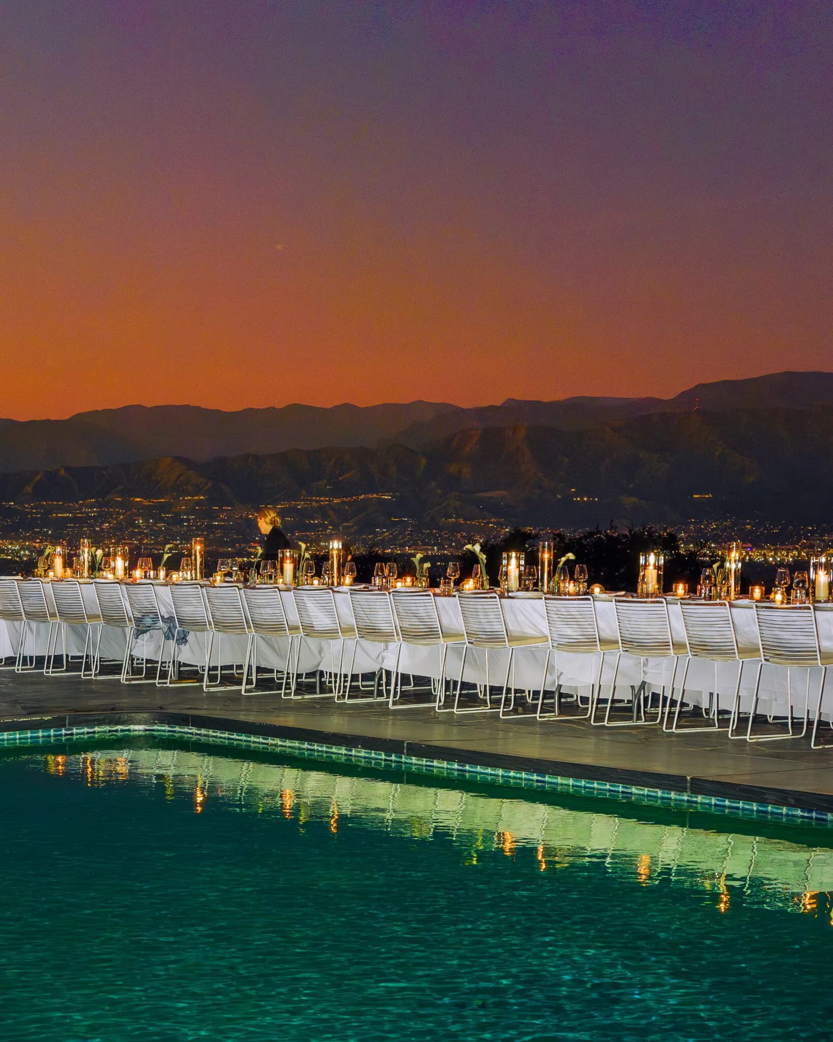 Outdoor event venue at dusk with rows of white chairs overlooking an illuminated pool and city skyline at night by Rachel Cope.