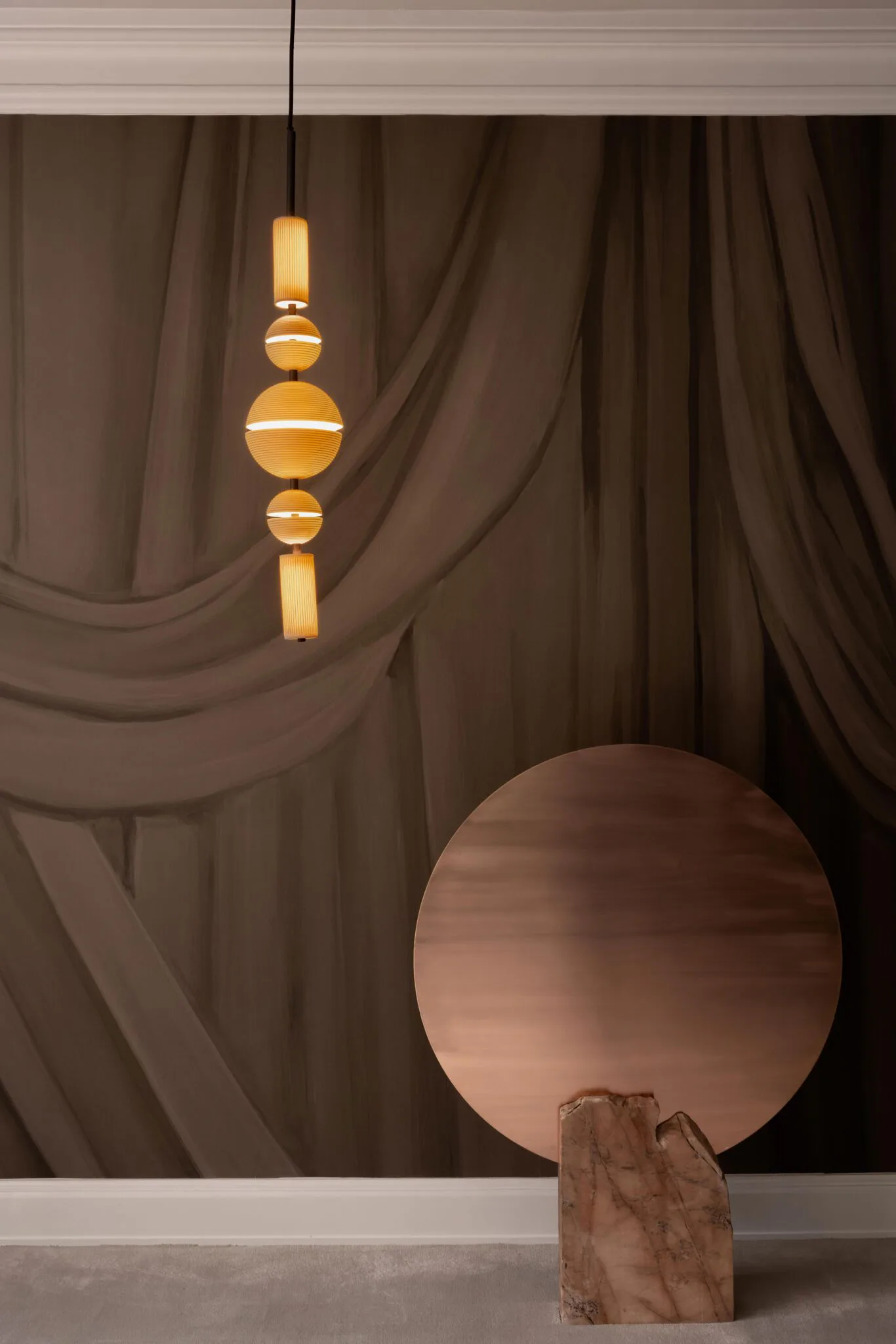 Warm-toned interior with draped brown curtains, geometric wooden sculpture with circular mirror, and sculptural brass pendant light by Rachel Cope.
