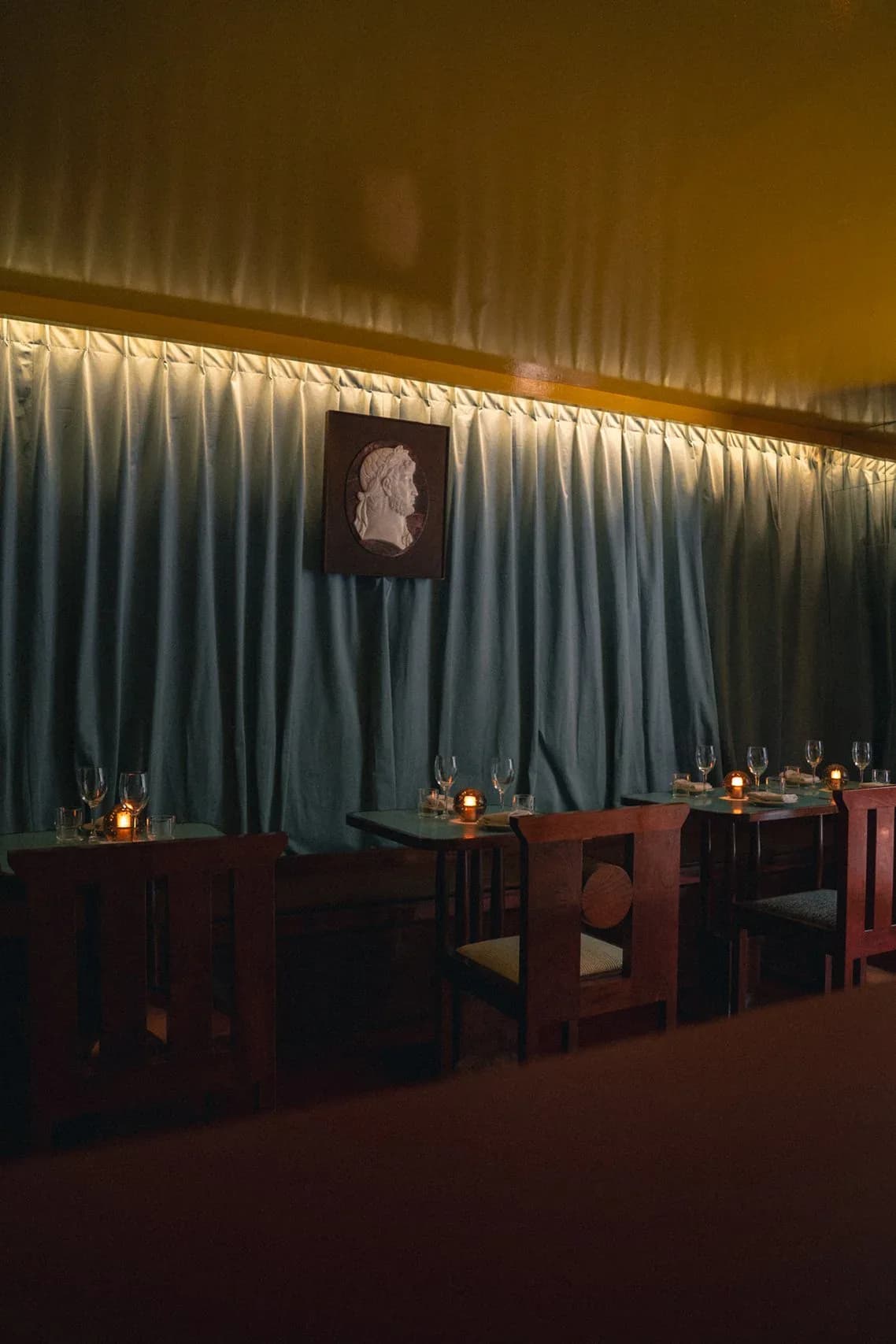 Atmospheric dining space with pleated blue-grey curtain backdrop, wooden tables and chairs, candlelit ambiance, and classical bust portrait by Rafael Prieto.