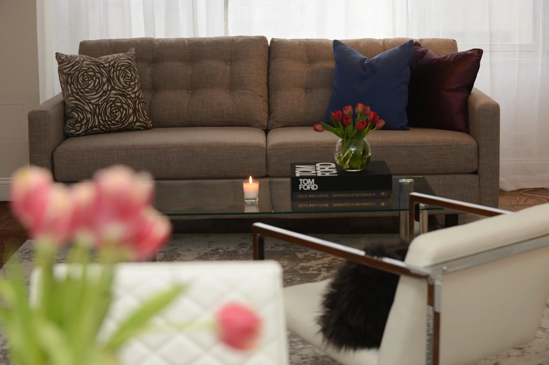 Modern living room with taupe sectional sofa, decorative pillows, glass coffee table with flowers, and neutral palette by Rahbia Allen.