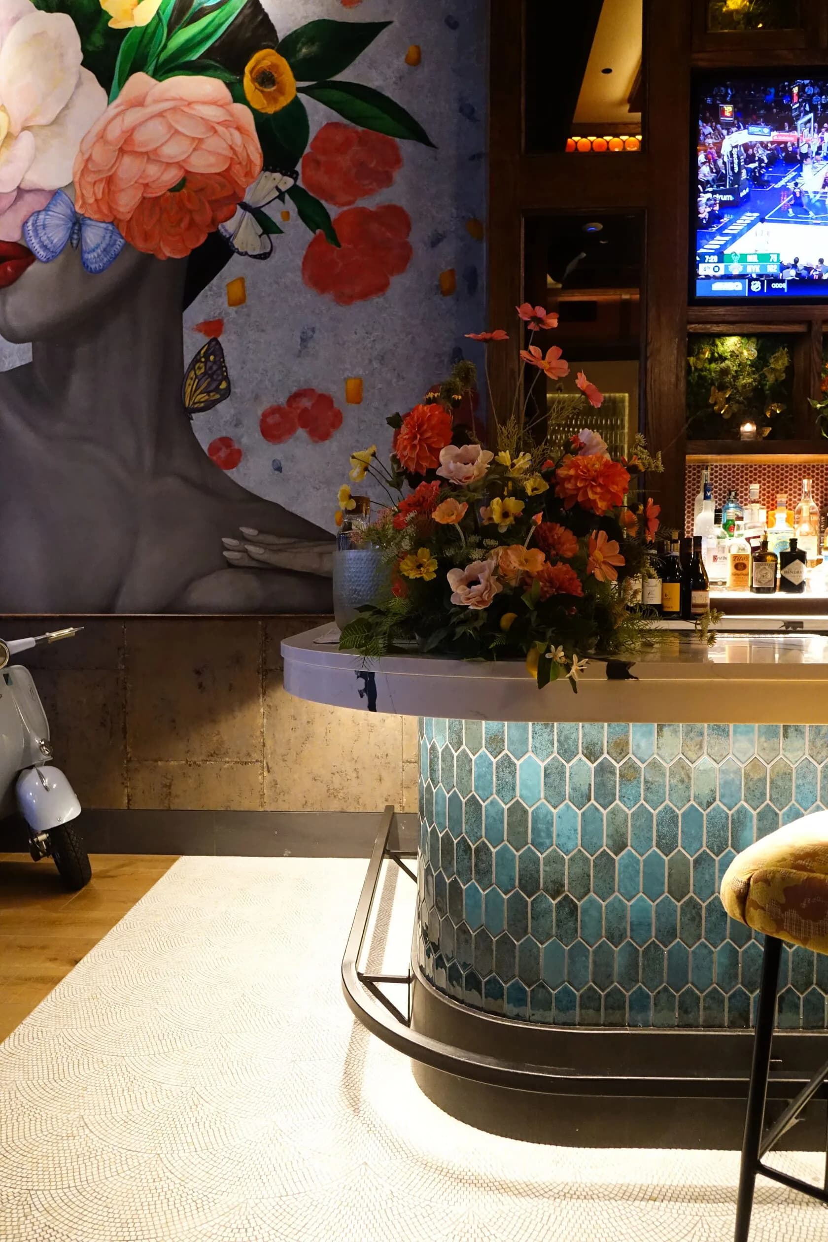 A modern bar or restaurant interior featuring a curved teal patterned counter, floral wall mural with coral and white flowers, and ambient lighting by Rddhima Golyan.